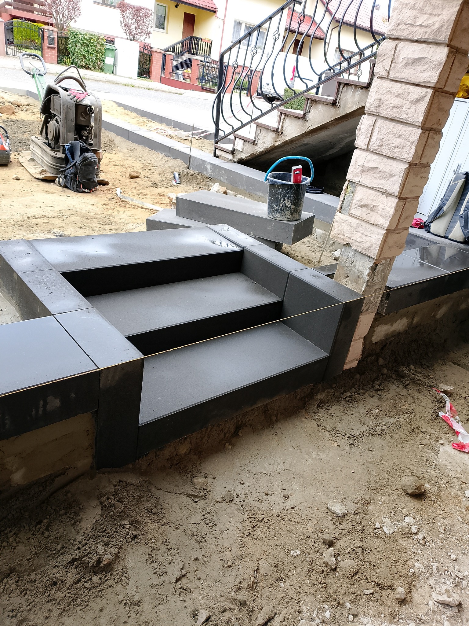 Montaż czarnych płyt na schodach wejściowych. Widoczny ubijak w tle. Etap prac brukarskich na posesji.