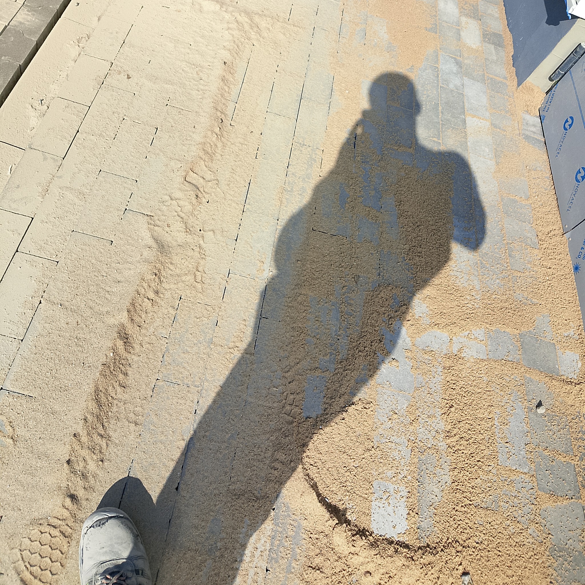 Świeżo ułożona kostka brukowa zasypana piaskiem, z widocznym cieniem osoby robiącej zdjęcie i fragmentem buta w lewym dolnym rogu.