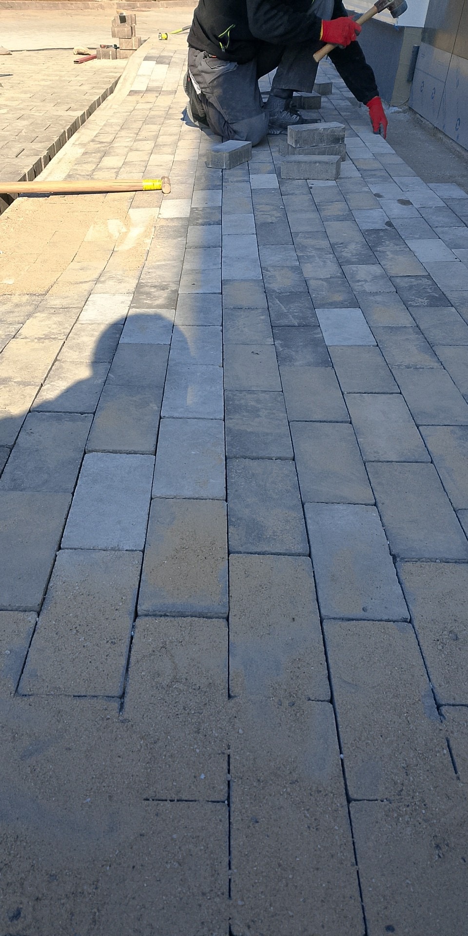 Praca brukarza w toku: układanie szarej kostki brukowej. Widoczny cień osoby, młotek i piasek między kostkami. Kąt ujęcia z góry, skupienie na detalu kostki.