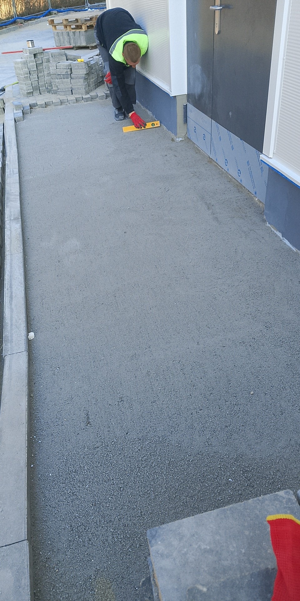 Pracownik w czerwonych rękawicach poziomuje podsypkę pod kostkę brukową przy budynku. Kostka ułożona wzdłuż ściany i krawężnika. Przygotowane materiały brukarskie.