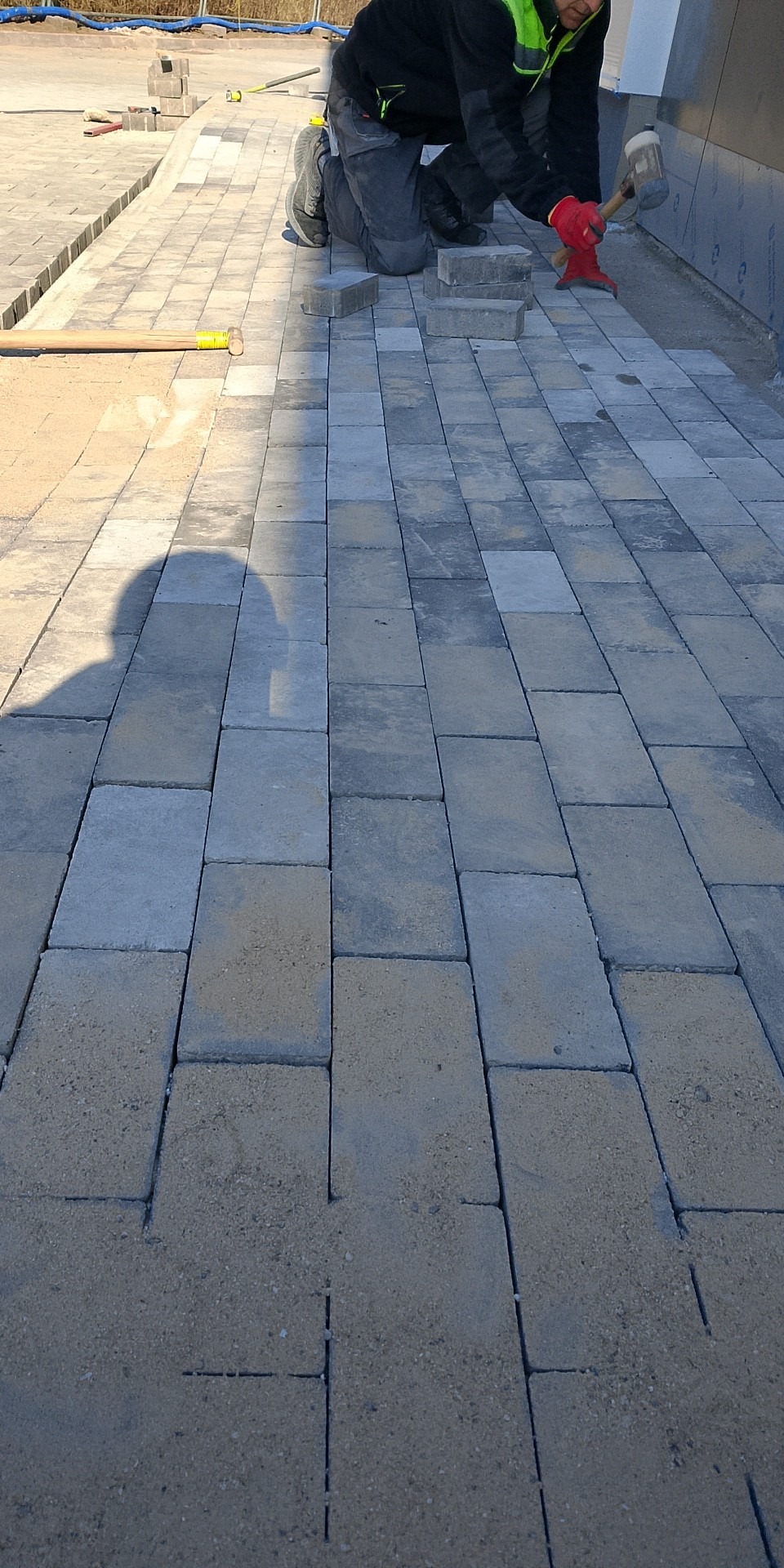 Układanie prostokątnej, szarej kostki brukowej. Brukarz w czerwonych rękawicach dobija kostkę gumowym młotkiem. Widoczny piasek i cień osoby fotografującej.