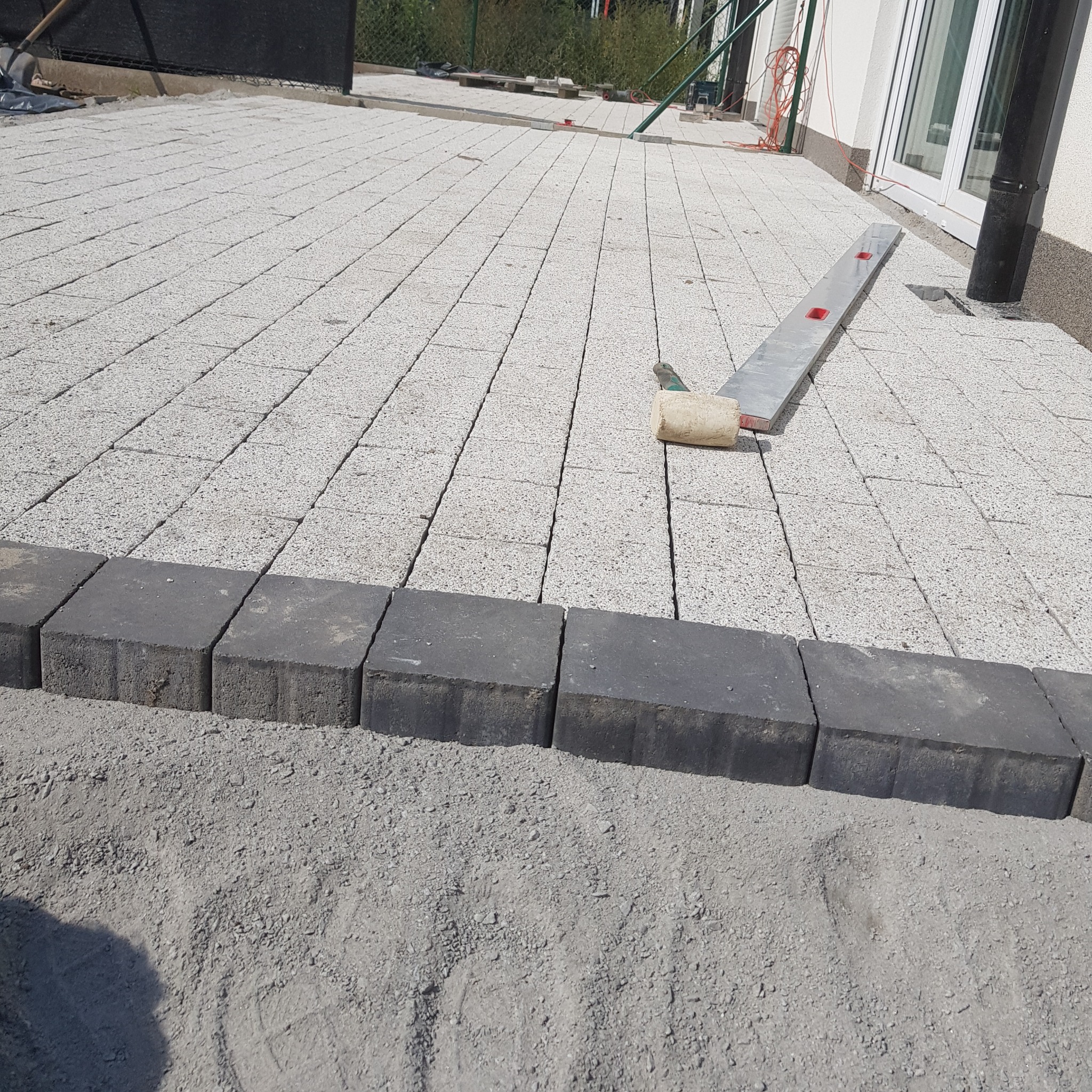 Jasnoszara kostka brukowa ułożona na posesji, obrzeża z ciemnej kostki, poziomica i gumowy młotek na powierzchni. Widok z góry na fragment nowej nawierzchni.
