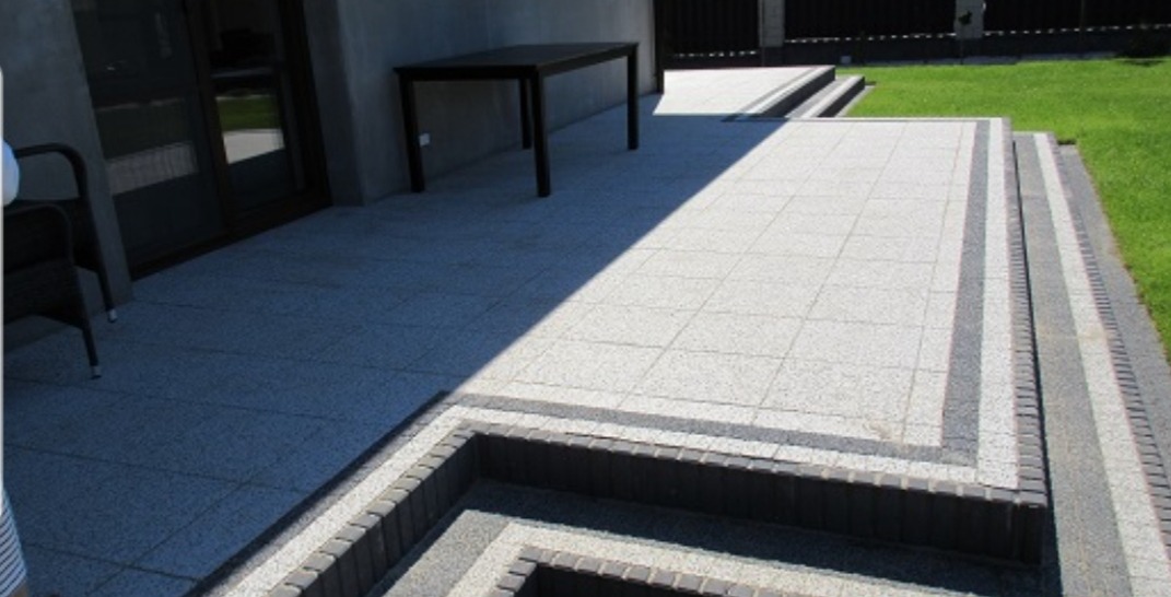 Ułożona kostka brukowa tworząca taras z niskimi schodami, obrzeża z ciemniejszej kostki, stół na tarasie, trawnik w tle, nowoczesny styl.