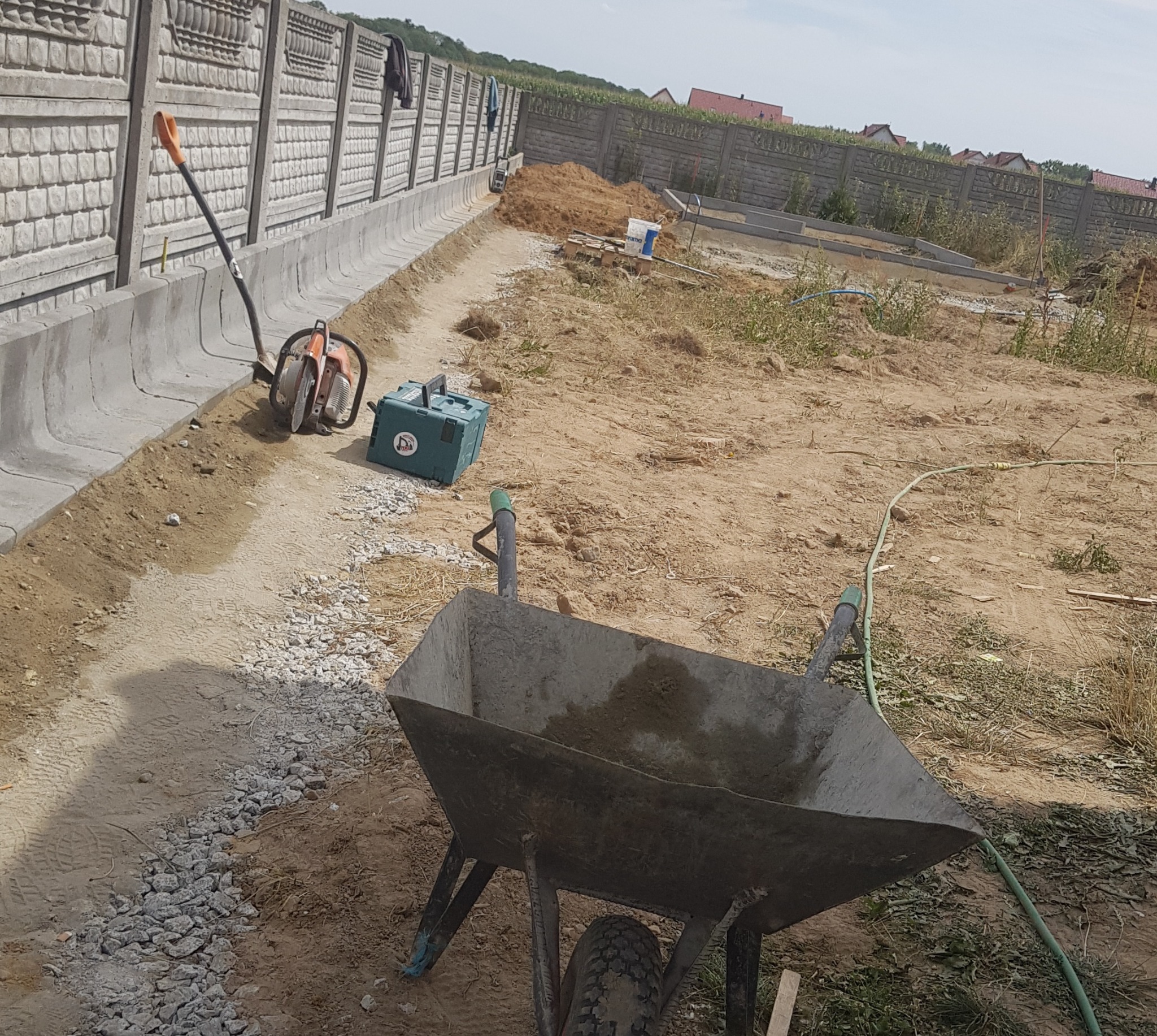 Budowa ogrodzenia panelowego z podmurówką prefabrykowaną. Widoczna taczka, piła do betonu i narzędzia. W tle domy i drzewa.