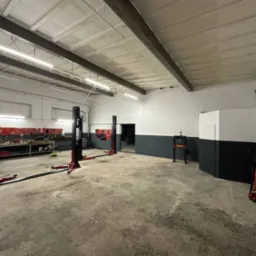 Przestronny warsztat samochodowy z trzema podnośnikami dwukolumnowymi i stanowiskiem narzędziowym. Betonowa podłoga i ściany pomalowane do połowy na szaro.