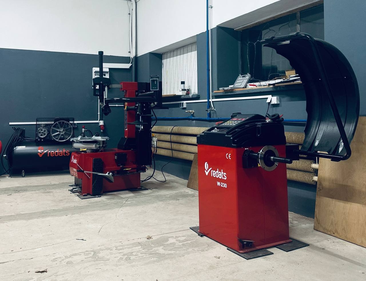 Wyważarka i montażownica do kół w warsztacie, marki Redats, model W-230. Widoczne okno i rury na ścianie. Posadzka betonowa z delikatnymi śladami użytkowania.