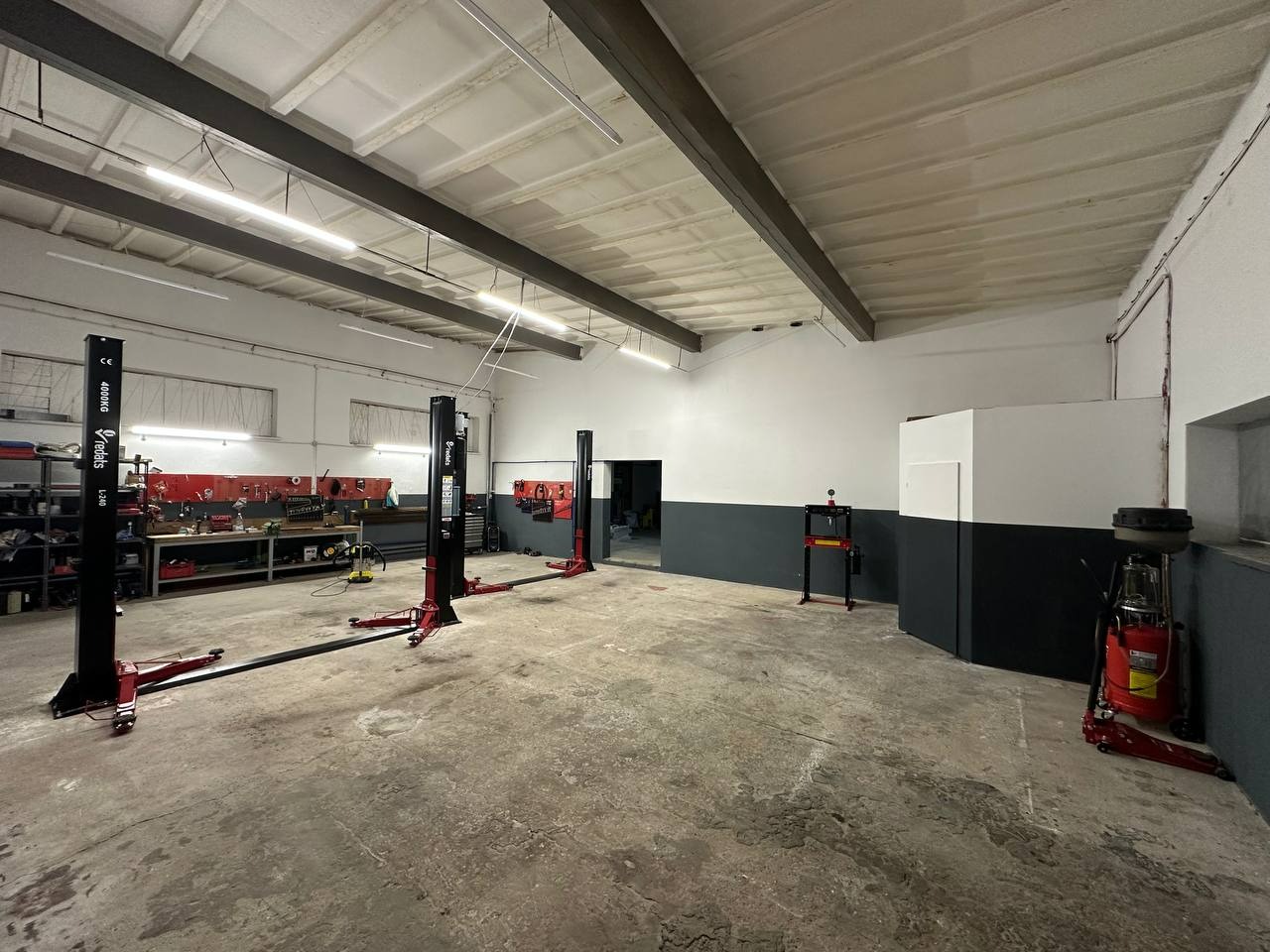 Przestronny warsztat samochodowy z trzema podnośnikami dwukolumnowymi i stanowiskiem narzędziowym. Betonowa podłoga i ściany pomalowane do połowy na szaro.