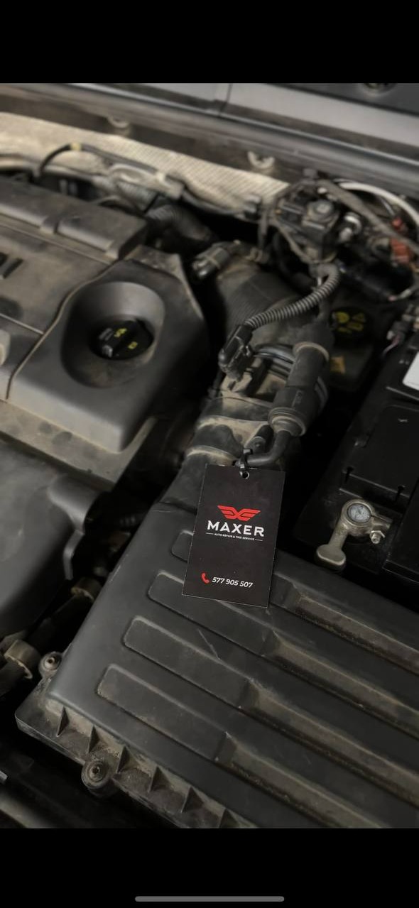 Silnik samochodowy z zawieszką reklamową MAXER. Widoczne elementy silnika, akumulator. Zawieszka z logo i numerem telefonu. Detal z warsztatu samochodowego.