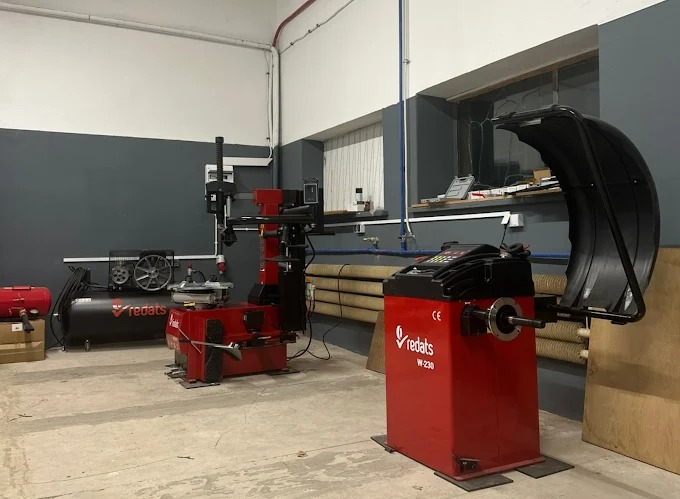 Wnętrze warsztatu z dwoma czerwonymi maszynami do obsługi opon, w tym wyważarką Redats W-230 i montażownicą. W tle widoczny kompresor i okno.