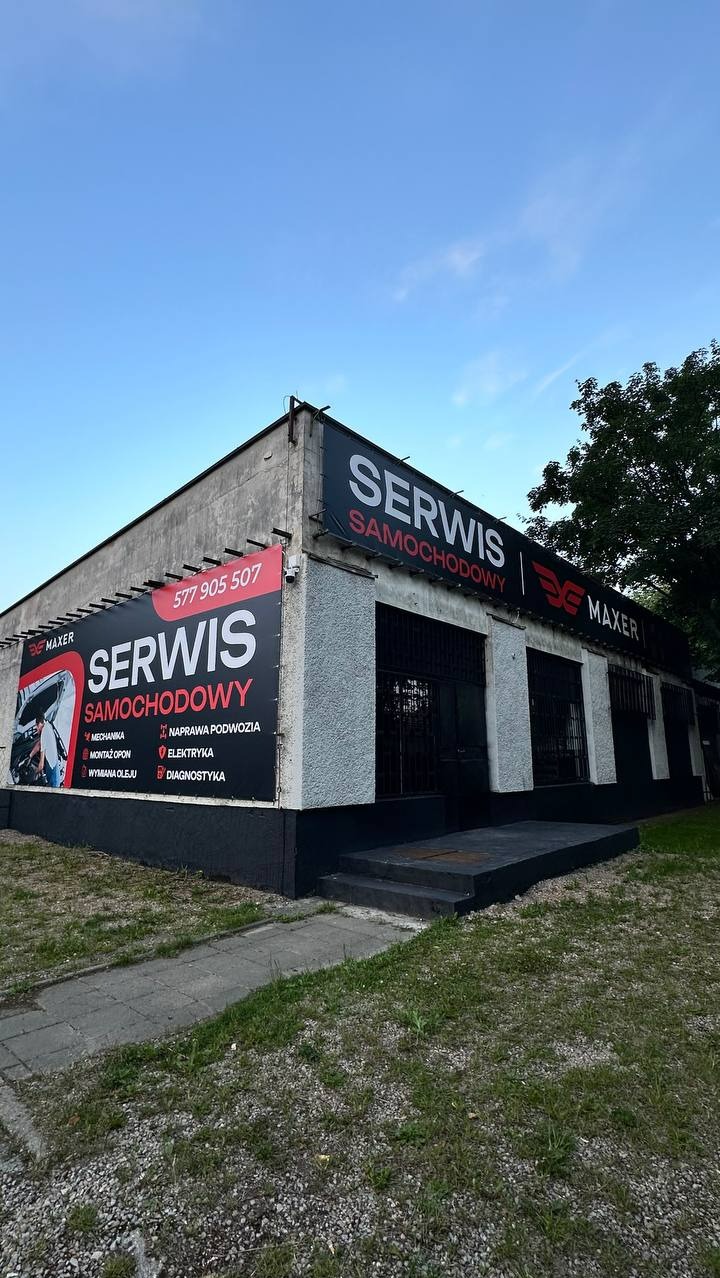 Budynek serwisu samochodowego z dużym banerem informacyjnym, na którym widnieją oferowane usługi: mechanika, elektryka, diagnostyka. Ujęcie z zewnątrz, w dzień.