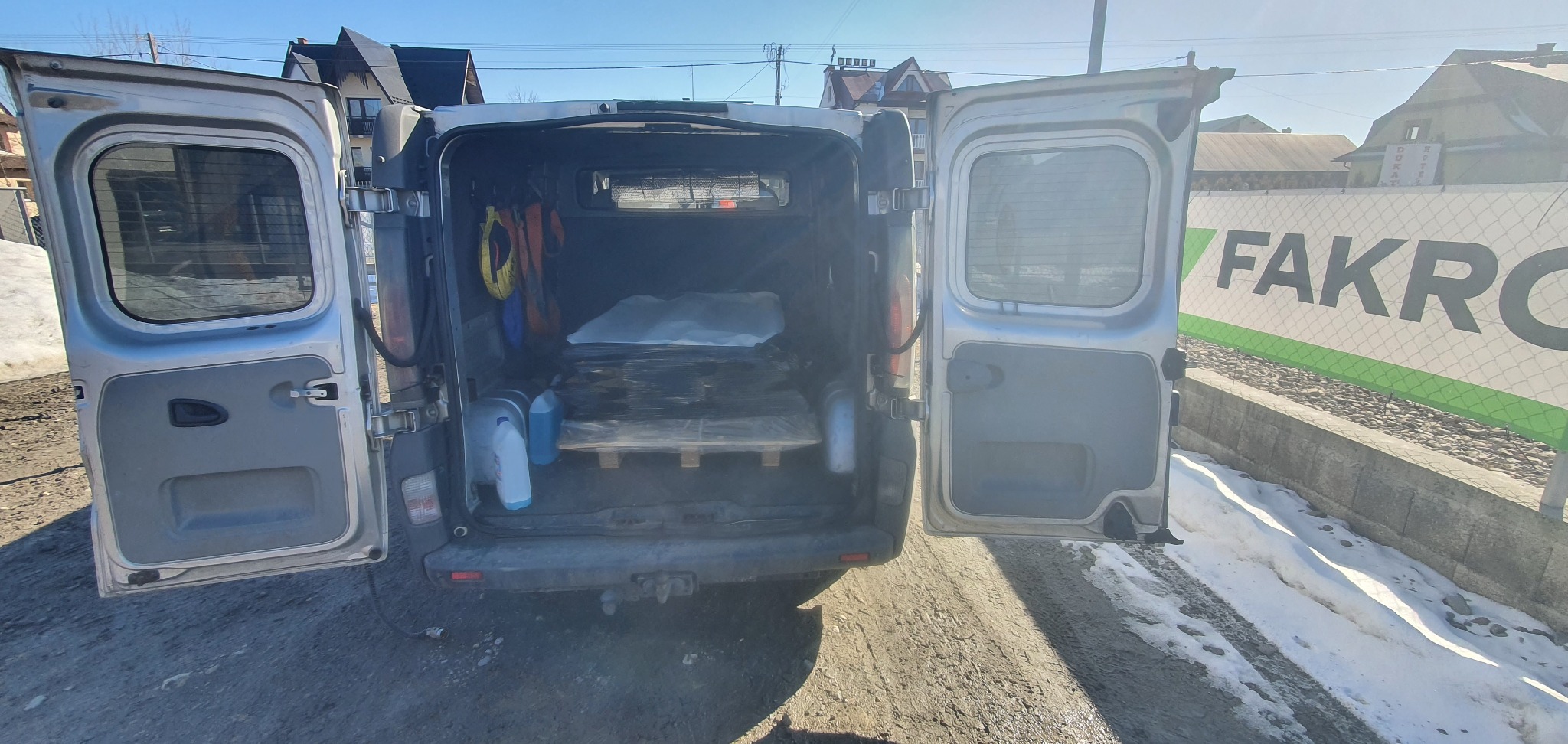 Srebrny bus z otwartymi drzwiami bagażnika, wewnątrz paleta z towarem. Widoczny baner reklamowy Fakro i fragment zabudowań w tle. Słońce oświetla teren.