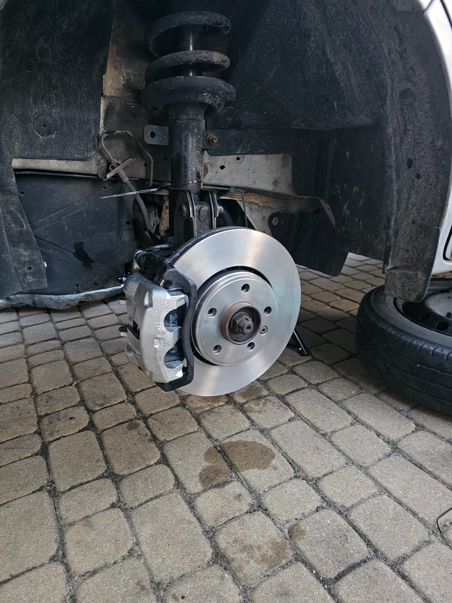Wymiana tarczy hamulcowej i klocków w samochodzie. Widoczny nowy, srebrny zacisk i tarcza hamulcowa. Obok leży zdjęte koło. Praca wykonana na kostce brukowej.