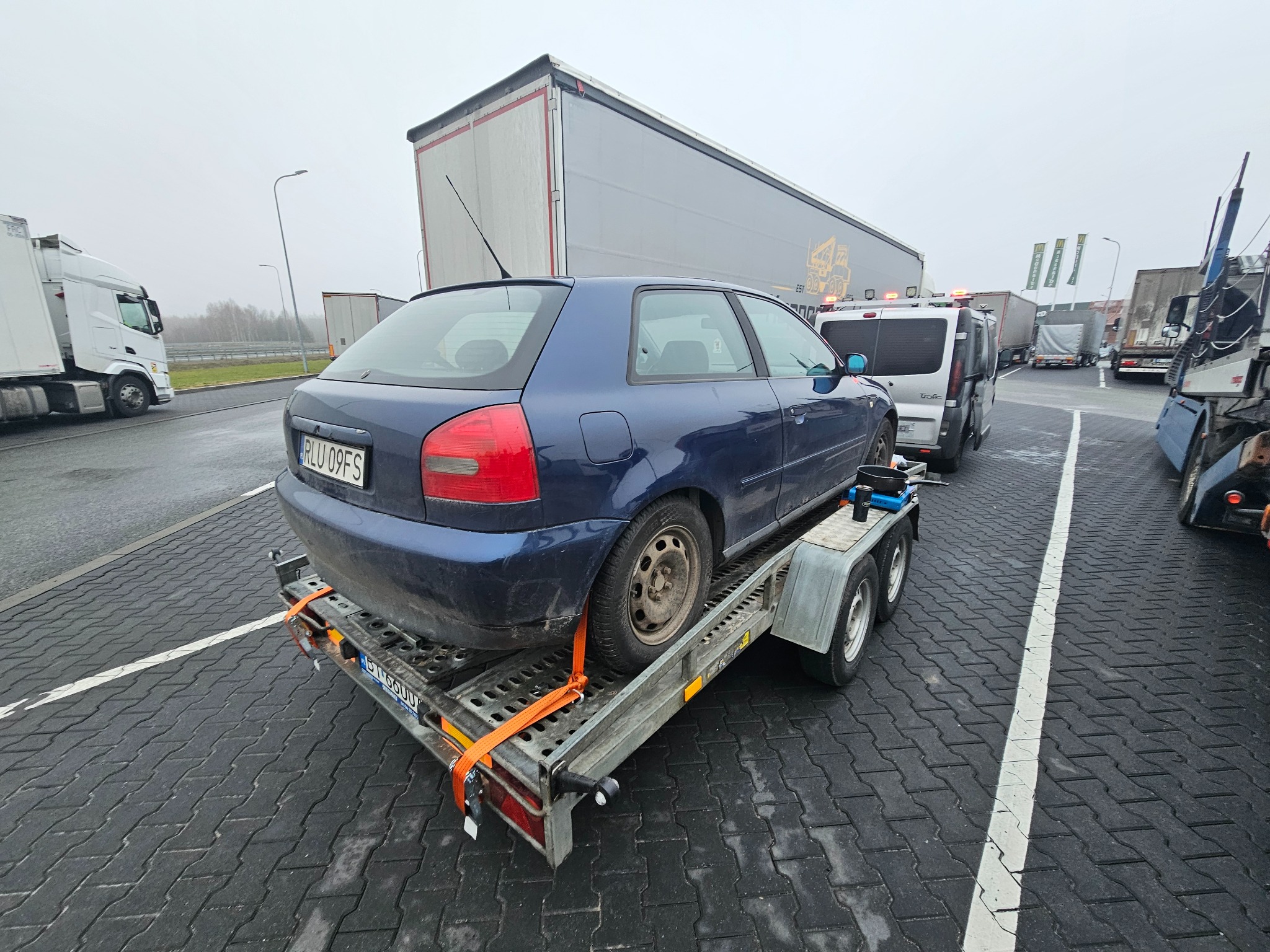 Niebieski samochód osobowy na niskopodwoziowej lawecie, zabezpieczony pomarańczowymi pasami transportowymi. W tle inne pojazdy ciężarowe i osobowe.