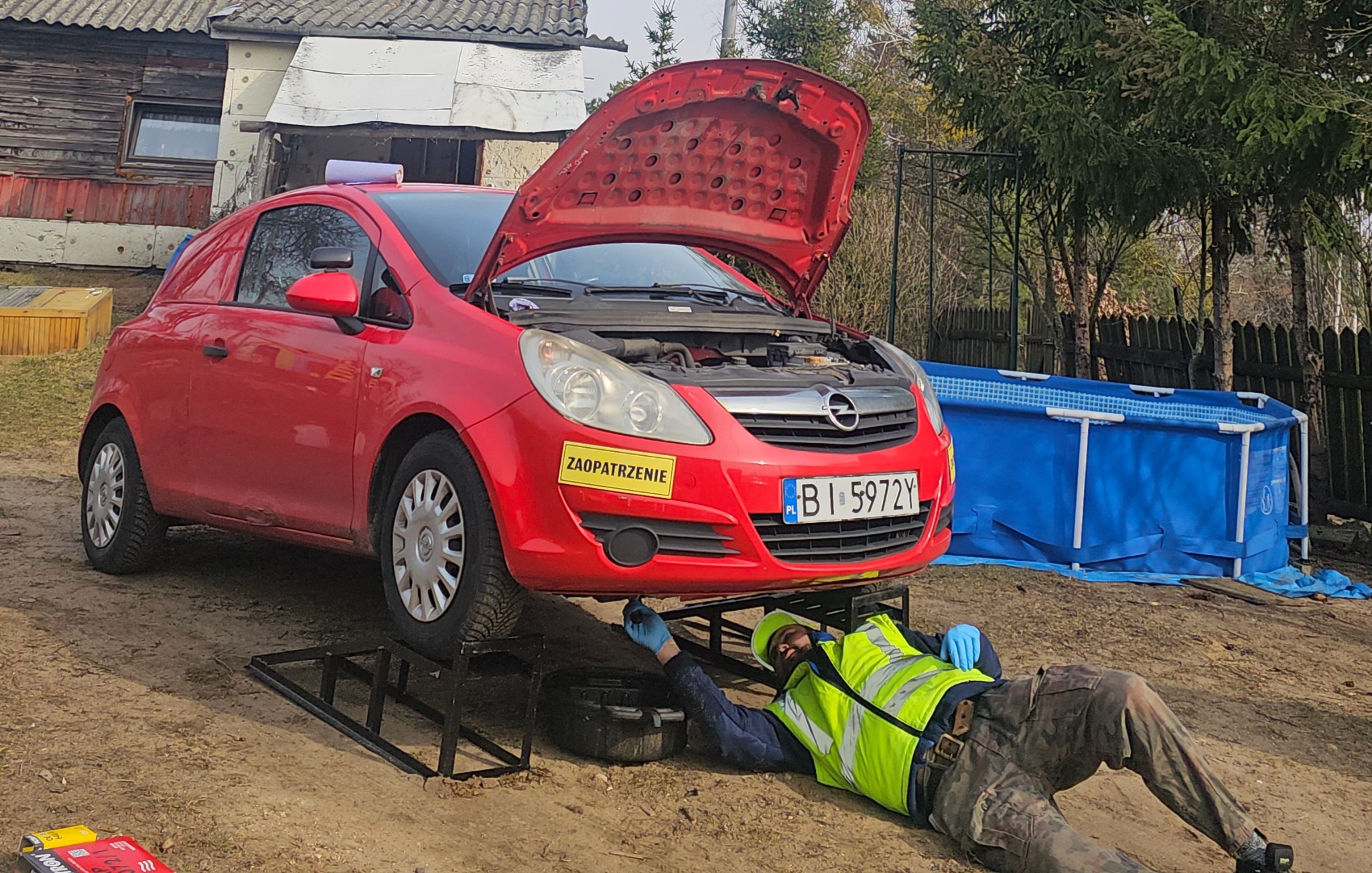 Czerwona Opel Corsa z otwartą maską, pod którą leży mechanik w kamizelce odblaskowej, dokonujący naprawy. W tle dom i basen.