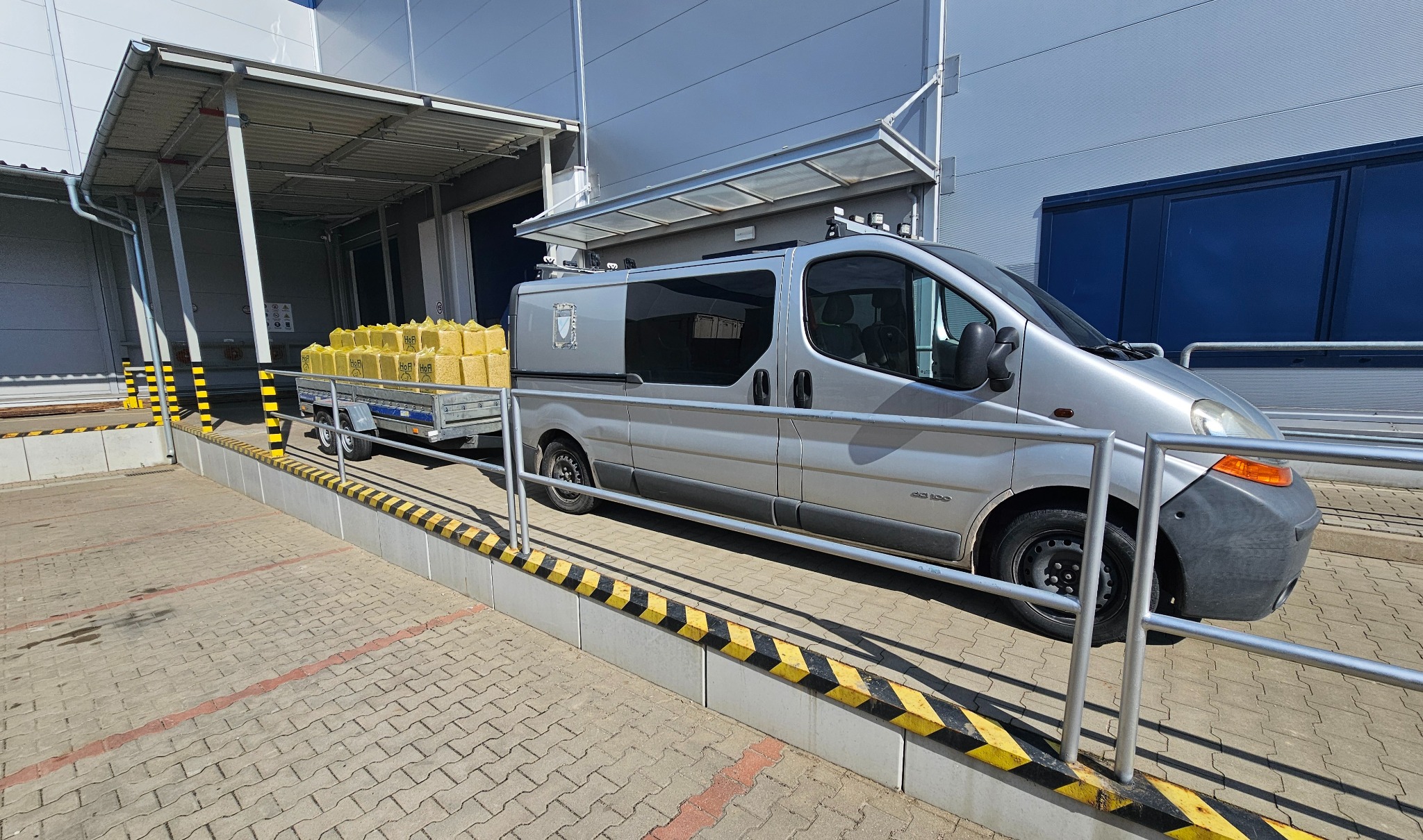 Srebrny bus z przyczepką wypełnioną żółtymi workami, zaparkowany przy rampie załadunkowej z żółto-czarnymi pasami ostrzegawczymi. Słoneczny dzień.