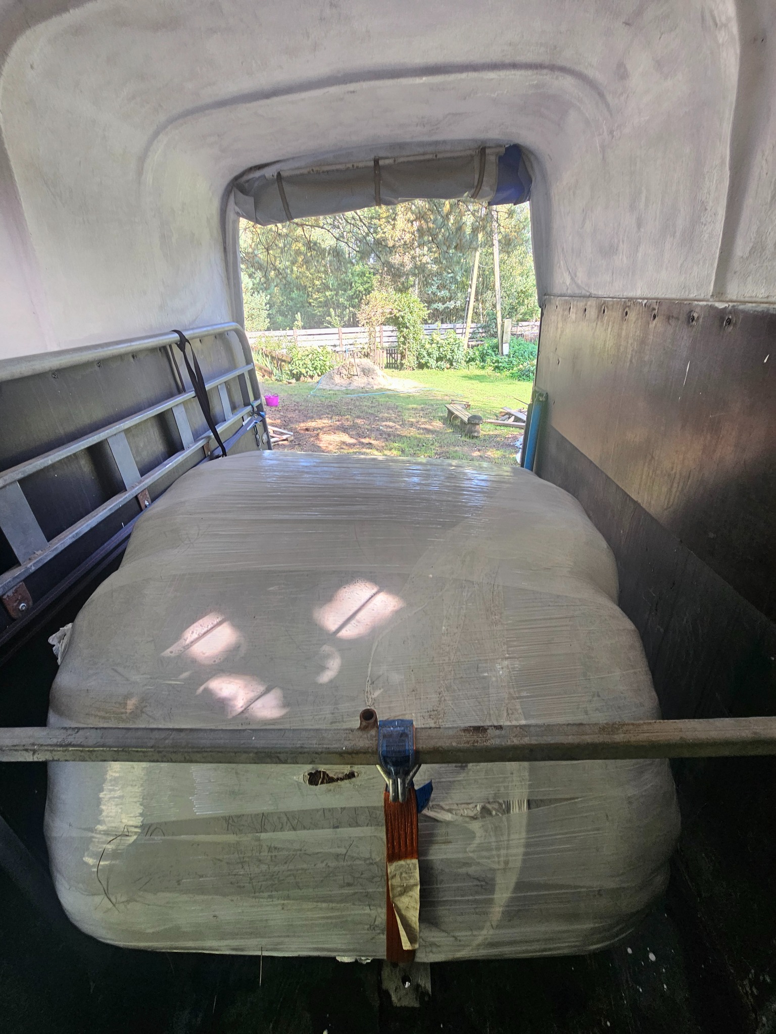 Ładunek owinięty folią zabezpieczającą, umieszczony w przestrzeni ładunkowej pojazdu transportowego. Widoczne elementy mocujące i fragment otoczenia przez otwarte drzwi.