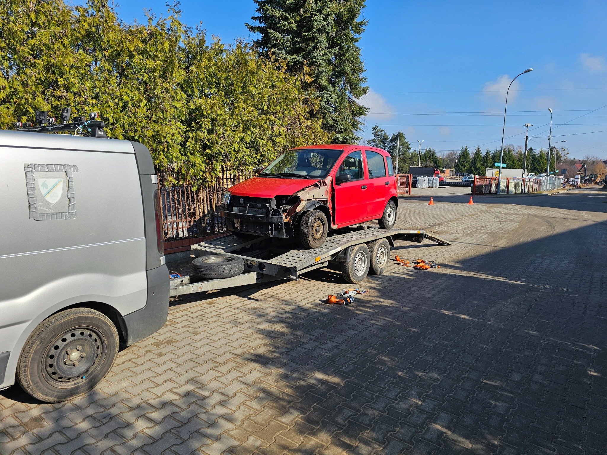 Laweta z uszkodzonym czerwonym autem osobowym na platformie, widok z boku, w tle zieleń i błękitne niebo. Auto bez przedniego zderzaka i reflektorów.