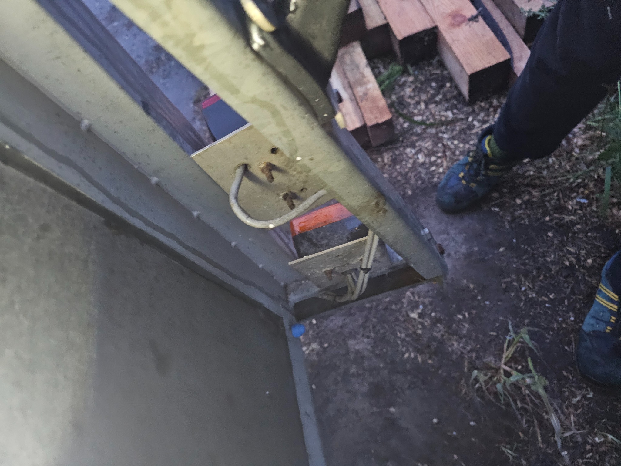 Fragment podwozia lawety z widocznymi elementami mocującymi i przewodami, obok leżą drewniane klocki i widać fragment buta osoby stojącej obok.