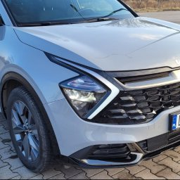 Atom Tomasz Kania - Szary SUV marki Kia Sportage z włączonymi światłami, zaparkowany na brukowanym placu. Widoczne detale: reflektory, atrapa chłodnicy i tablica rejestracyjna.