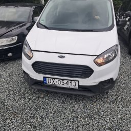 Atom Tomasz Kania - Biały Ford Transit Courier z polską tablicą rejestracyjną DX 05413, zaparkowany na żwirowym parkingu, widziany z przodu, obok czarnego auta.
