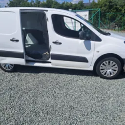 Biały bus dostawczy z otwartymi drzwiami, zaparkowany na żwirowym podłożu. Widoczny bok pojazdu i fragment zieleni w tle. Oświetlenie dzienne, słoneczne.