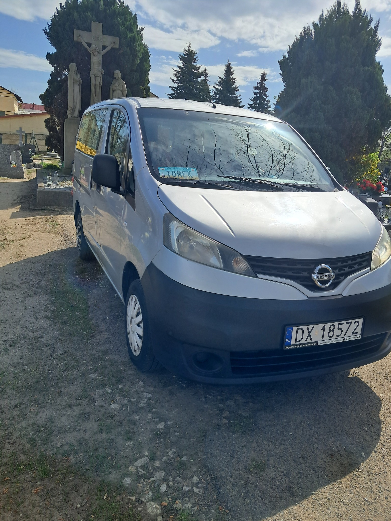 Srebrny Nissan NV200 na tle cmentarza z krzyżem i nagrobkami, tablica rejestracyjna z napisem 'Tomek' na przedniej szybie.