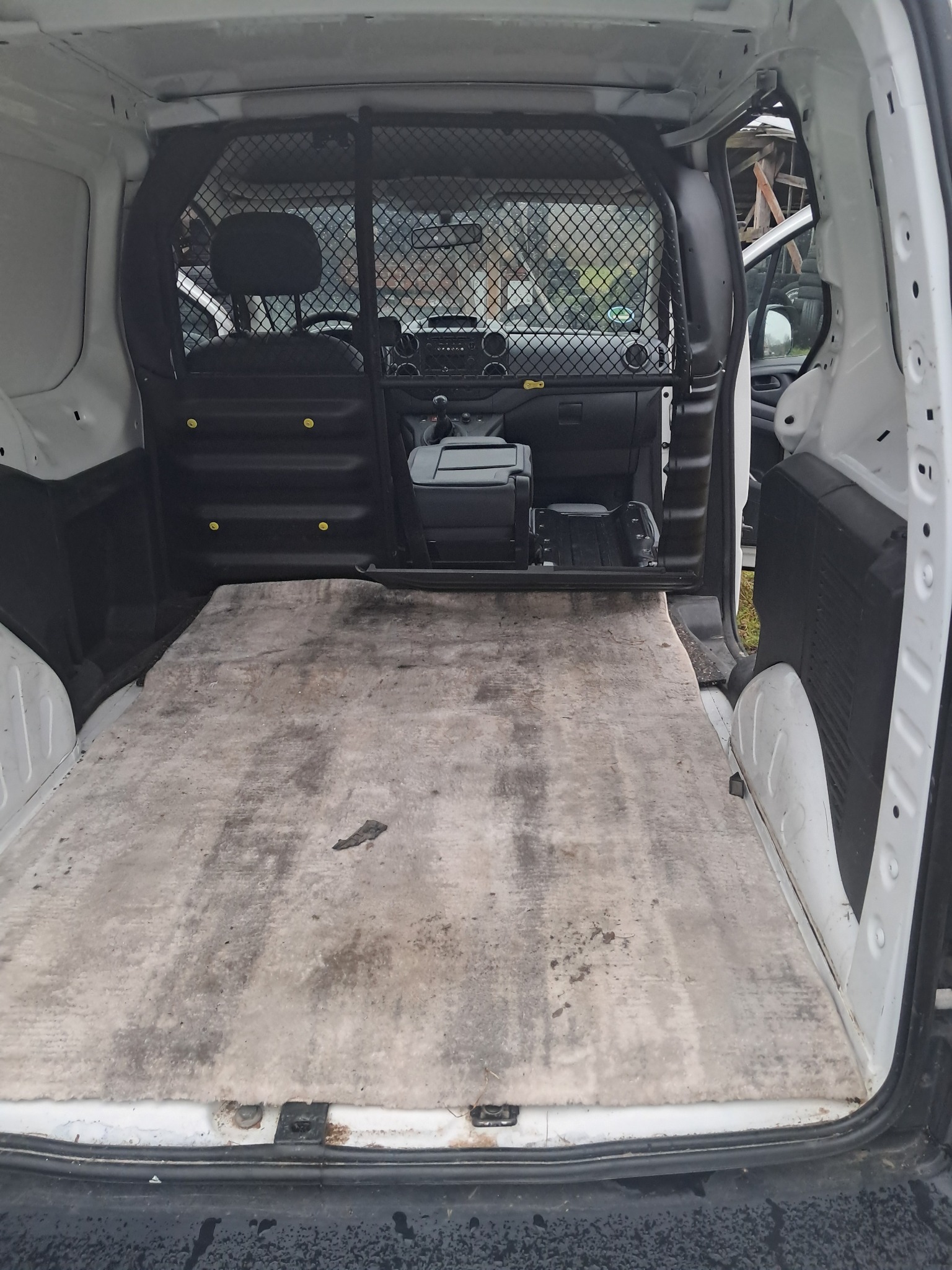 Wnętrze białego busa z kratą oddzielającą przestrzeń ładunkową. Widoczna brudna podłoga w przestrzeni ładunkowej i deska rozdzielcza z przodu.