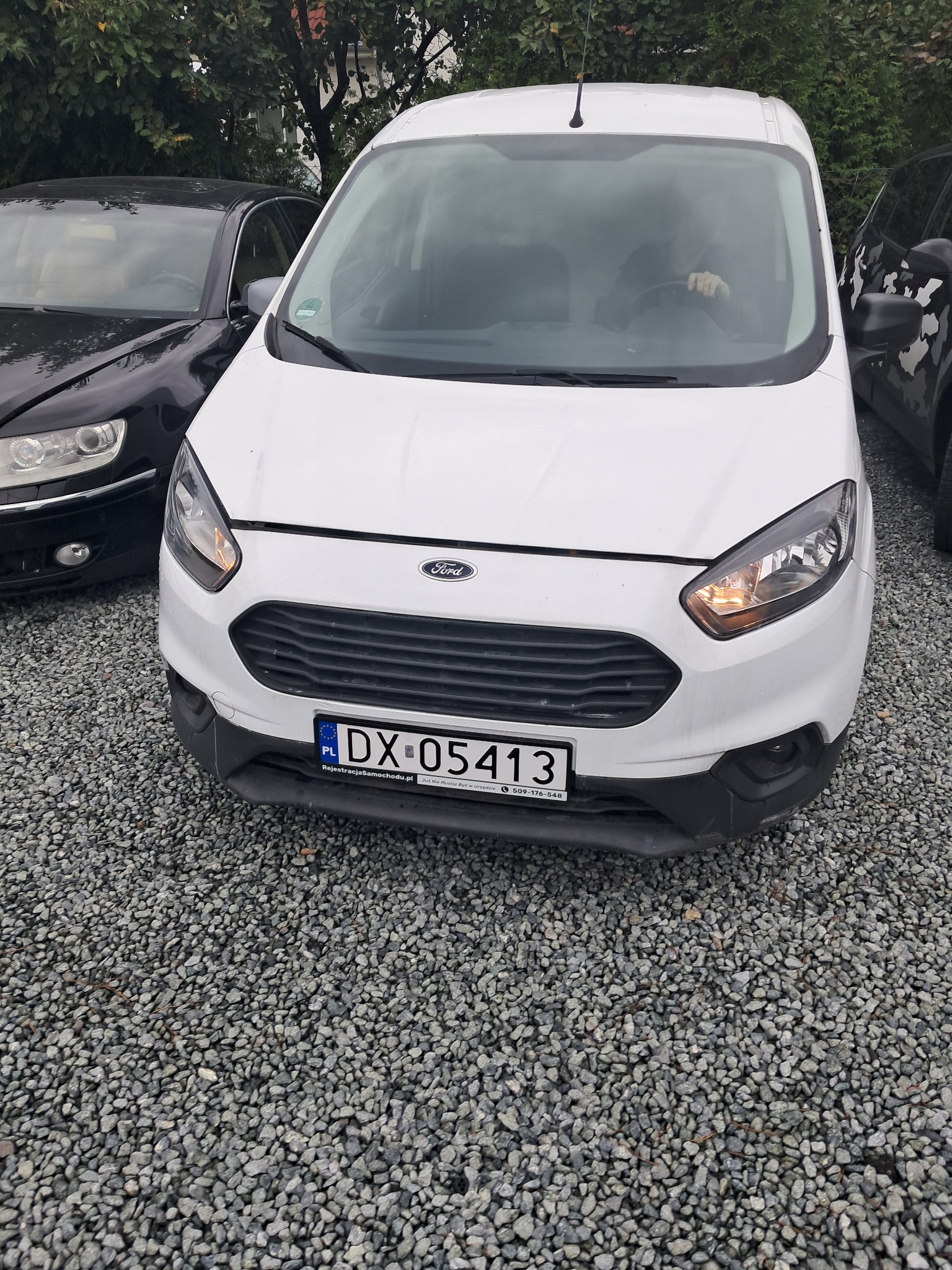 Biały Ford Transit Courier z polską tablicą rejestracyjną DX 05413, zaparkowany na żwirowym parkingu, widziany z przodu, obok czarnego auta.