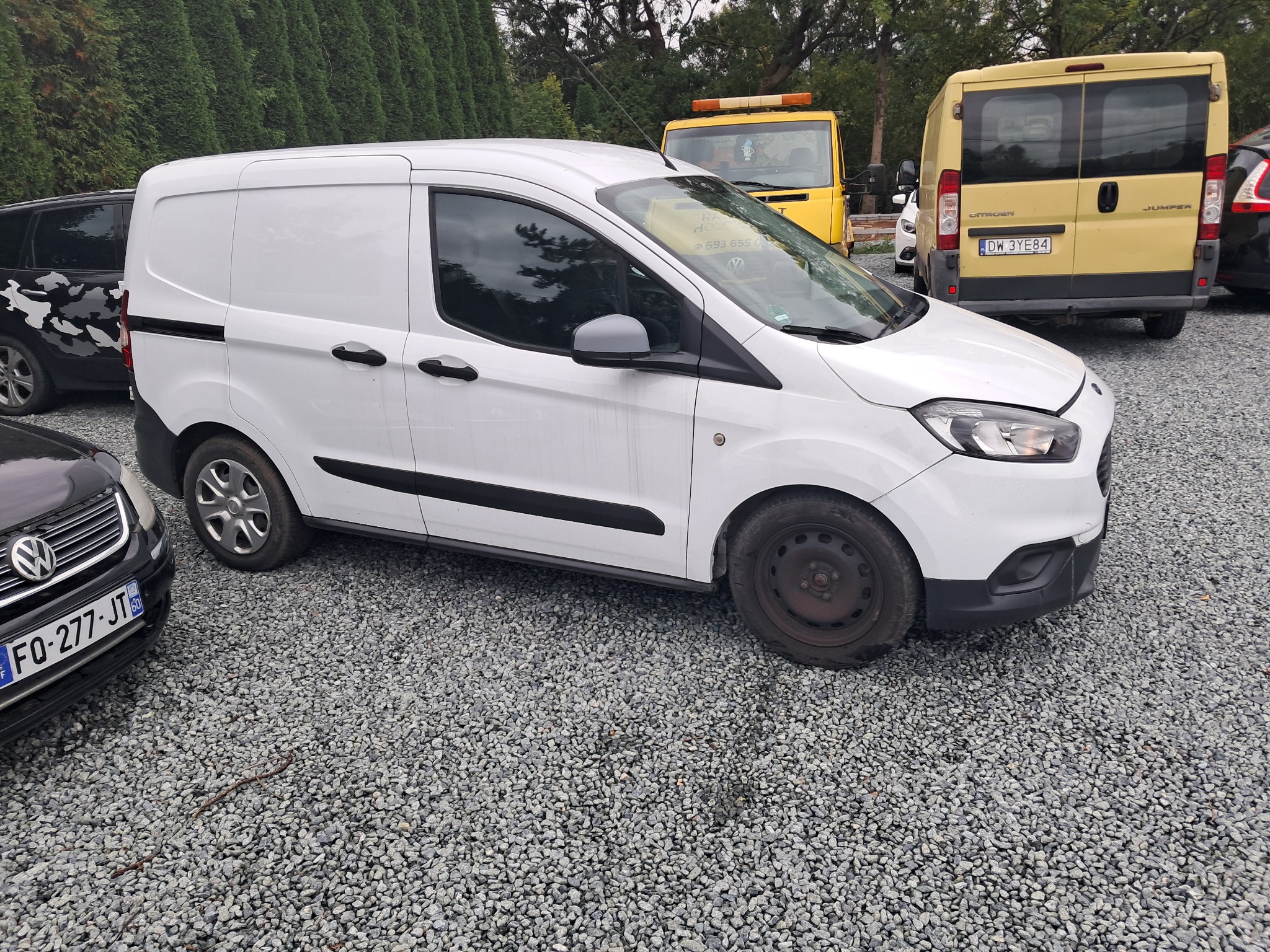 Biały bus Ford Transit Courier na żwirowym parkingu, z widoczną lawetą w tle. Pojazd z ciemnymi listwami i klamkami, bez reklamy, zaparkowany obok czarnego samochodu.
