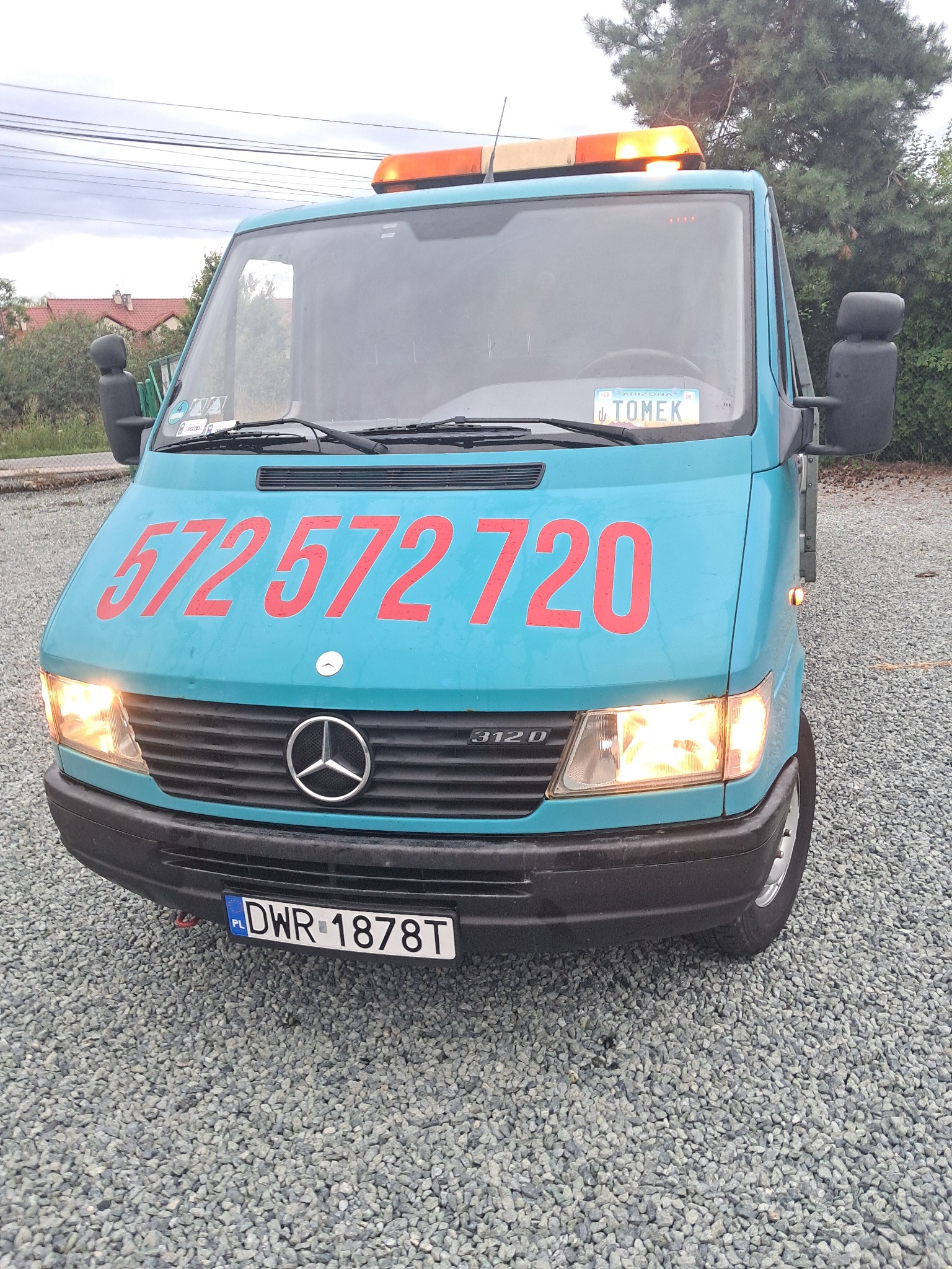 Błękitny Mercedes-Benz 312D z numerem telefonu na masce, pomarańczowym światłem ostrzegawczym i tablicą rejestracyjną DWR 1878T na żwirowym podłożu.