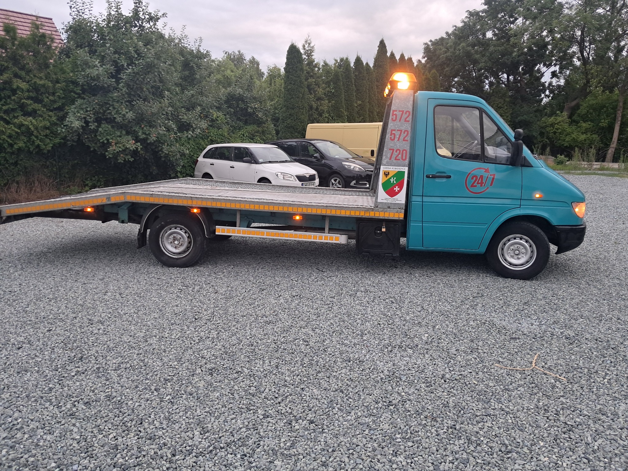 Turkusowa laweta z pomarańczowym światłem ostrzegawczym na żwirowym podłożu. Na lawecie dwa samochody osobowe. Widoczne numery kontaktowe na nadwoziu.