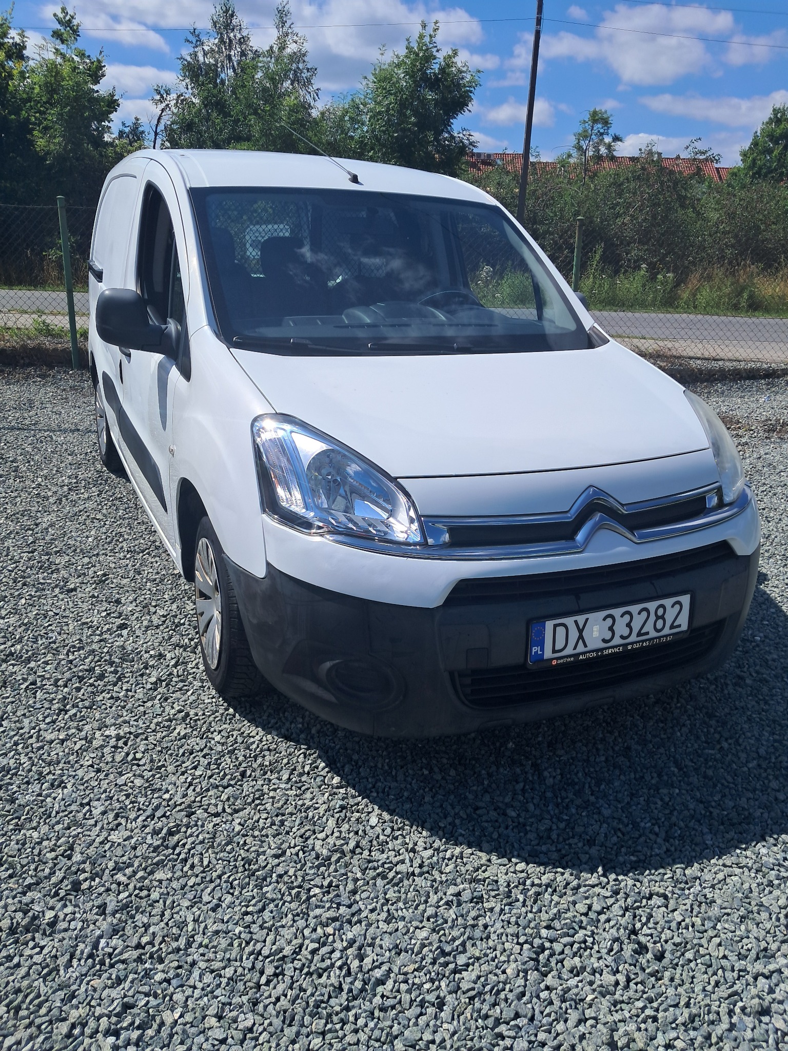 Biały bus marki Citroen Berlingo na żwirowym podłożu, z widoczną tablicą rejestracyjną i elementami krajobrazu w tle, w słoneczny dzień.