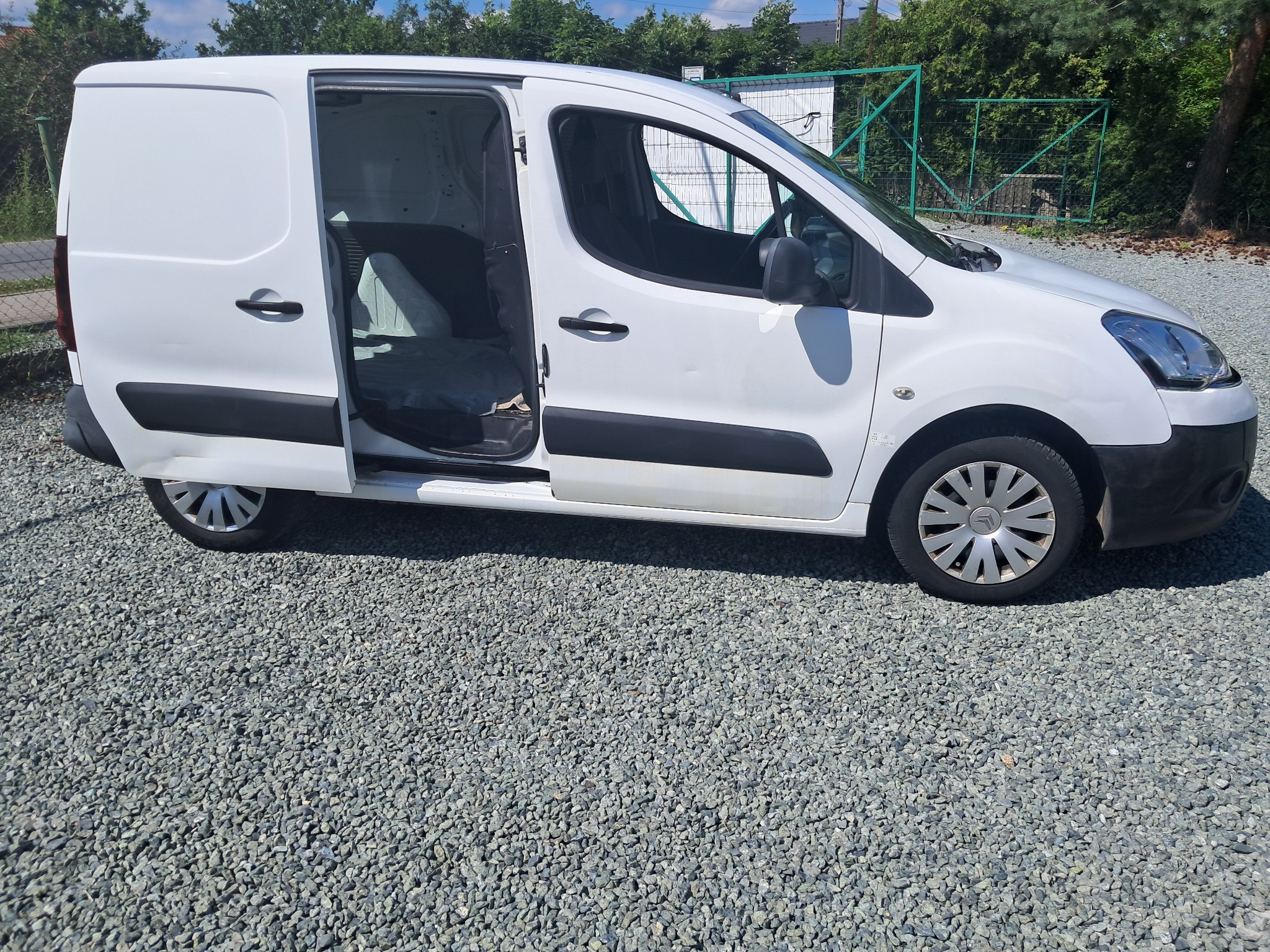 Biały bus dostawczy z otwartymi drzwiami, zaparkowany na żwirowym podłożu. Widoczny bok pojazdu i fragment zieleni w tle. Oświetlenie dzienne, słoneczne.