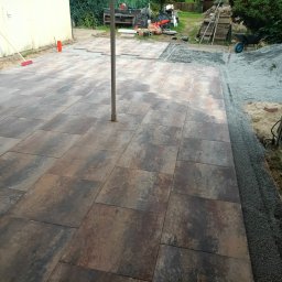 Lukk bud - Częściowo ułożona kostka brukowa na posesji. Widoczny fragment podwórka z przygotowanym podłożem i narzędziami. Kostka w odcieniach brązu i szarości.