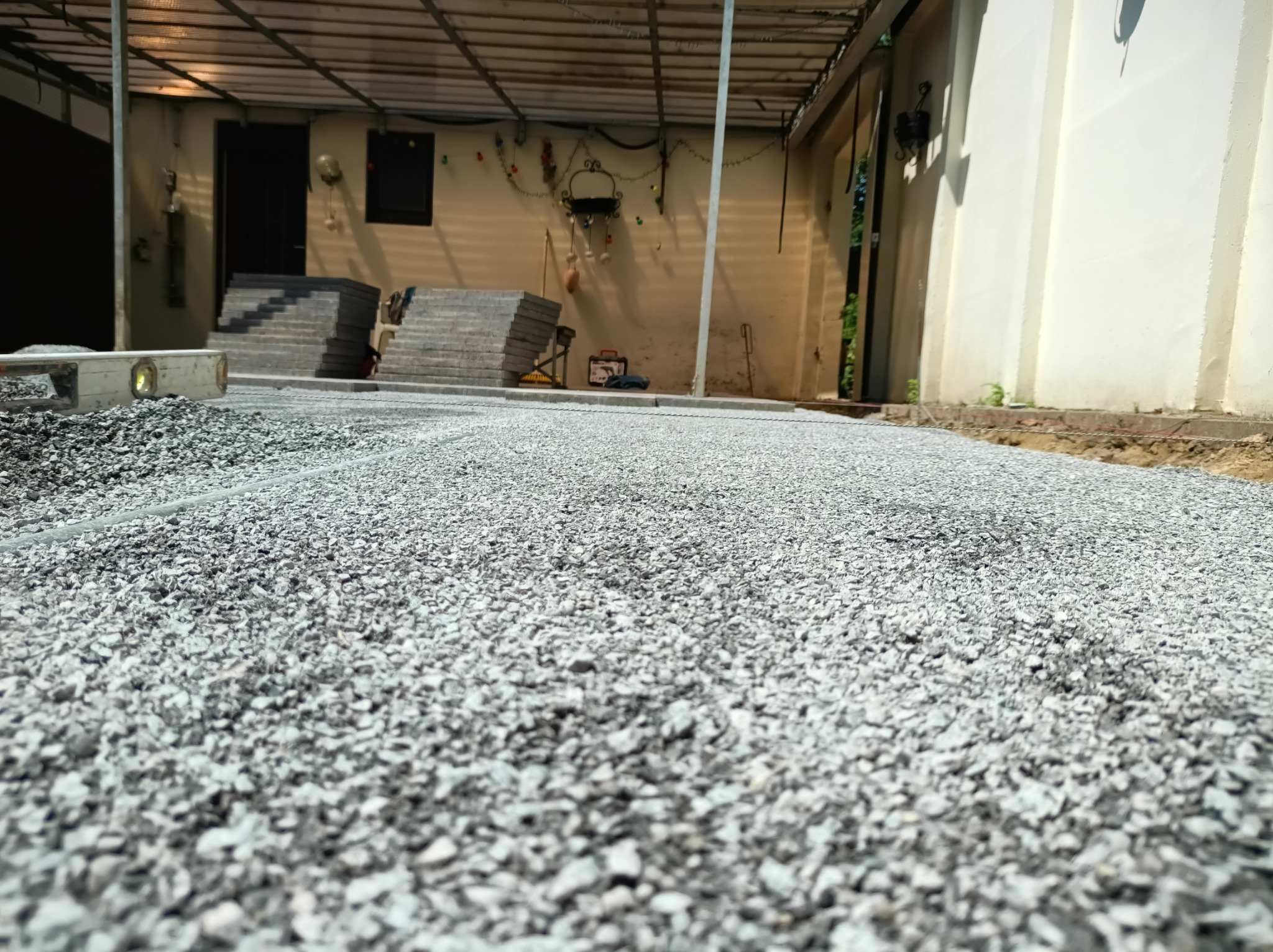 Przygotowanie podłoża z szarego kruszywa, widoczny poziomica i kostka. Prace brukarskie w toku, wyrównywanie terenu podłoża. Kąt z dołu, oświetlenie dzienne.