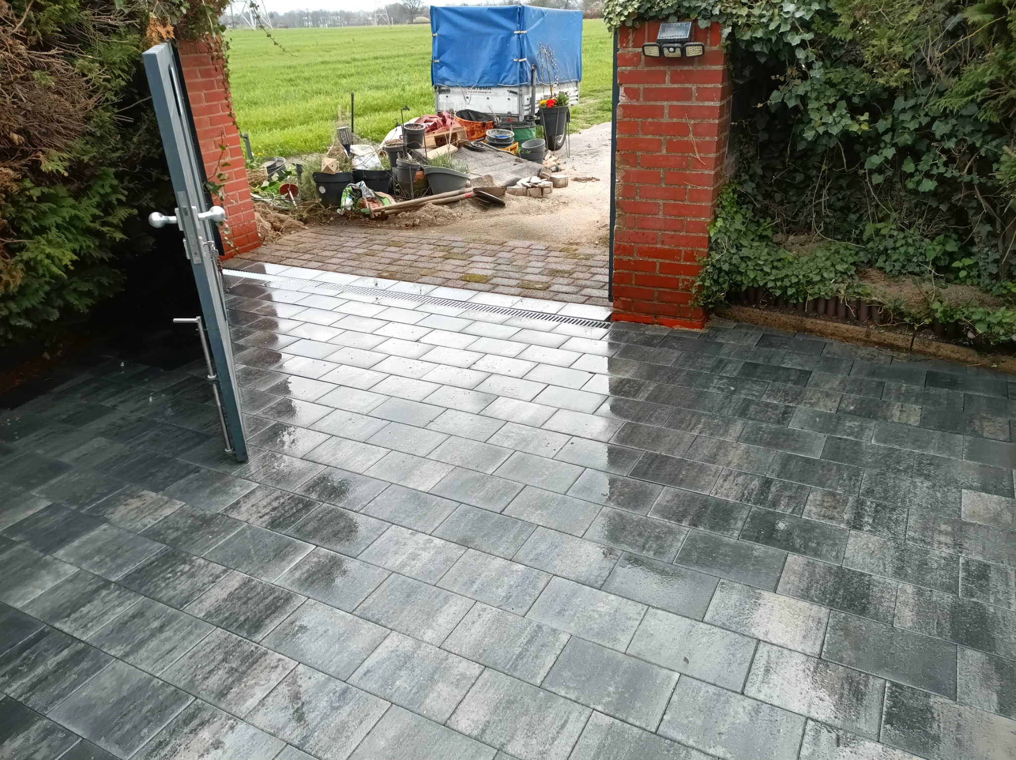 Świeżo ułożona, mokra kostka brukowa w odcieniach szarości, widziana pod kątem. W tle brama z cegły i widoczny krajobraz z przyczepą.