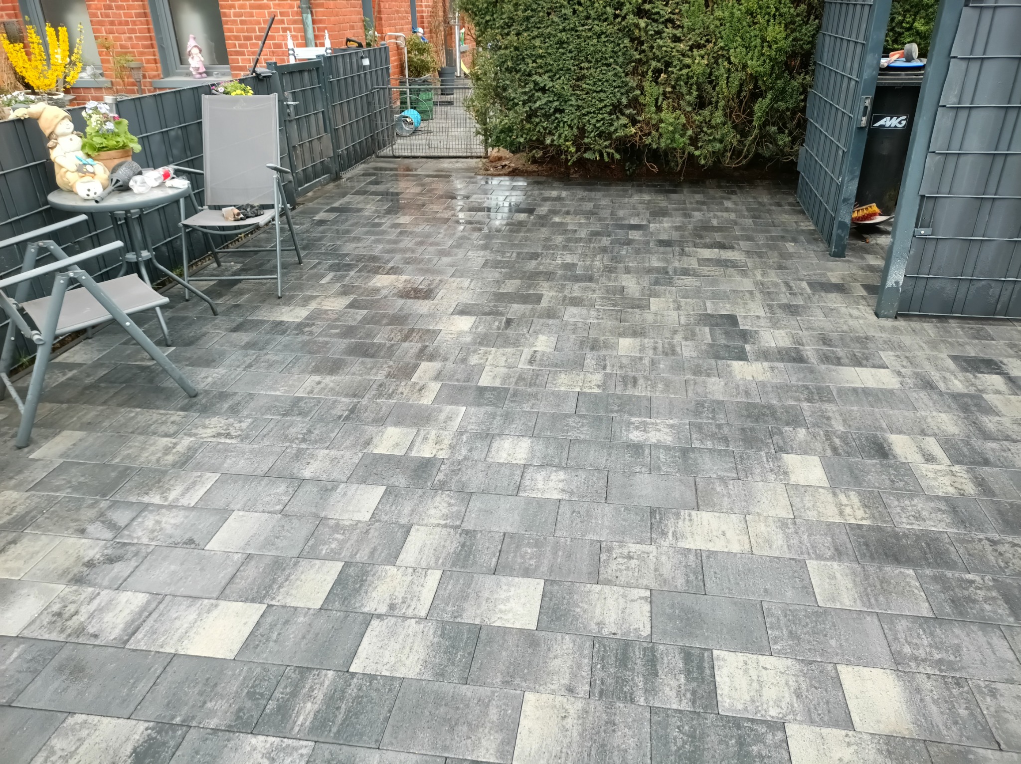 Ułożona, szara kostka brukowa na podwórku z szarą, metalową bramą i ogrodzeniem. Widoczne krzesła i stolik z dekoracjami. Częściowo widoczny budynek z czerwonej cegły.