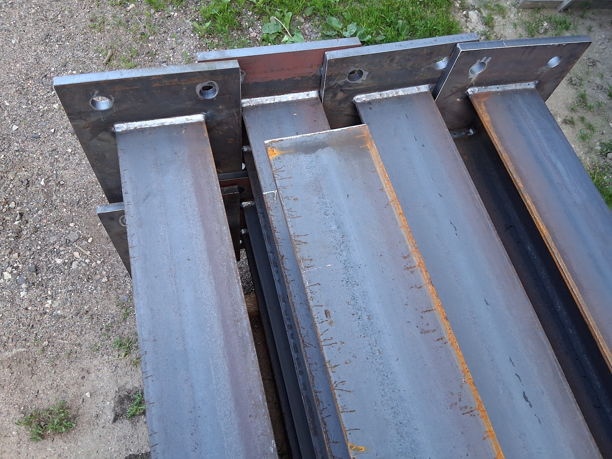 Stalowe, surowe słupy z przyspawanymi płytami bazowymi, z widocznymi otworami, ułożone na ziemi. Widok z góry, blik światła na krawędziach.