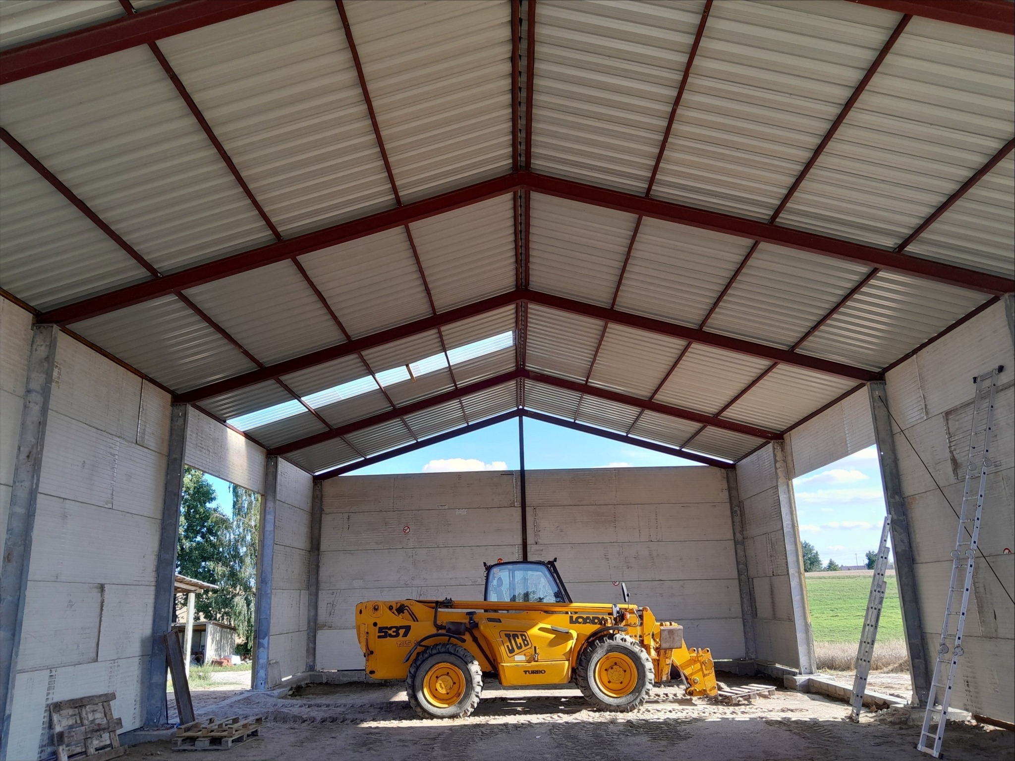 Żółta ładowarka teleskopowa JCB w surowej hali z betonowych płyt i stalowej konstrukcji dachu, z widocznymi drabinami i paletami. Przez otwartą ścianę widać krajobraz.