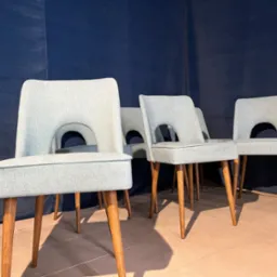 Ustawione w rzędzie krzesła tapicerowane w odcieniach szarości z drewnianymi nogami na tle ciemnoniebieskiej ściany. Widoczne charakterystyczne wycięcie w oparciu.