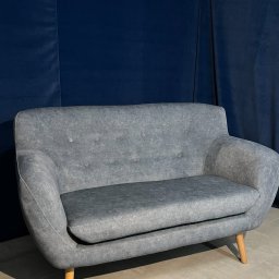 Odnova Meble Tapicerowane Wiktor Tomaszewski - Szara sofa dwuosobowa w stylu retro na drewnianych nóżkach, z tapicerowanymi guzikami na oparciu, na tle ciemnoniebieskiej, aksamitnej zasłony.