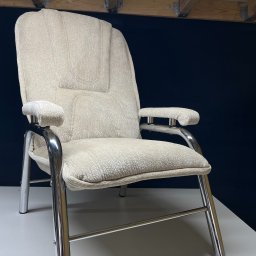 Odnova Meble Tapicerowane Wiktor Tomaszewski - Beżowy fotel z tapicerką o wyraźnej fakturze i chromowanym stelażu, ustawiony na białym blacie na tle ciemnego tła. Widoczne elementy konstrukcji drewnianej.