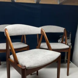 Trzy odnowione krzesła z drewnianą ramą i szarą tapicerką na siedziskach i oparciach, w tle narzędzie tapicerskie i rolka waty.