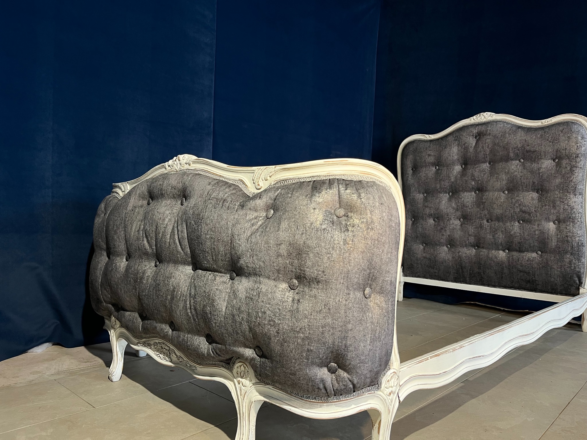 Stylowe, szare łóżko tapicerowane z białymi, zdobionymi ramami na tle granatowej ściany. Widoczny zagłówek i część ramy. Klasyczny design.