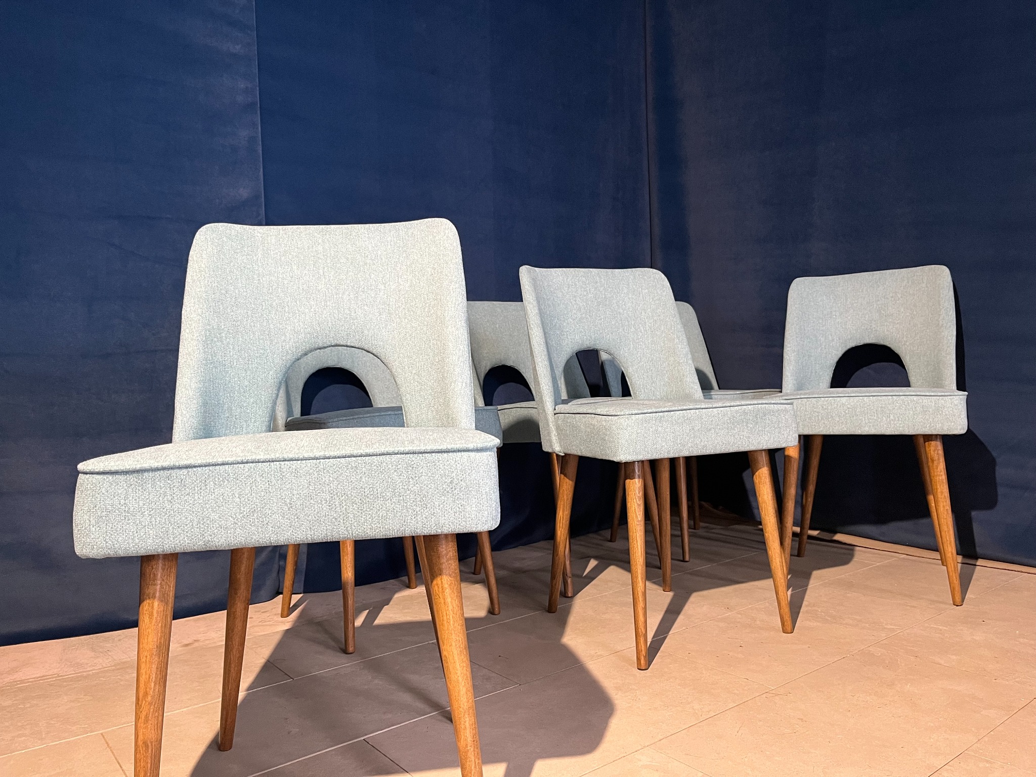 Ustawione w rzędzie krzesła tapicerowane w odcieniach szarości z drewnianymi nogami na tle ciemnoniebieskiej ściany. Widoczne charakterystyczne wycięcie w oparciu.