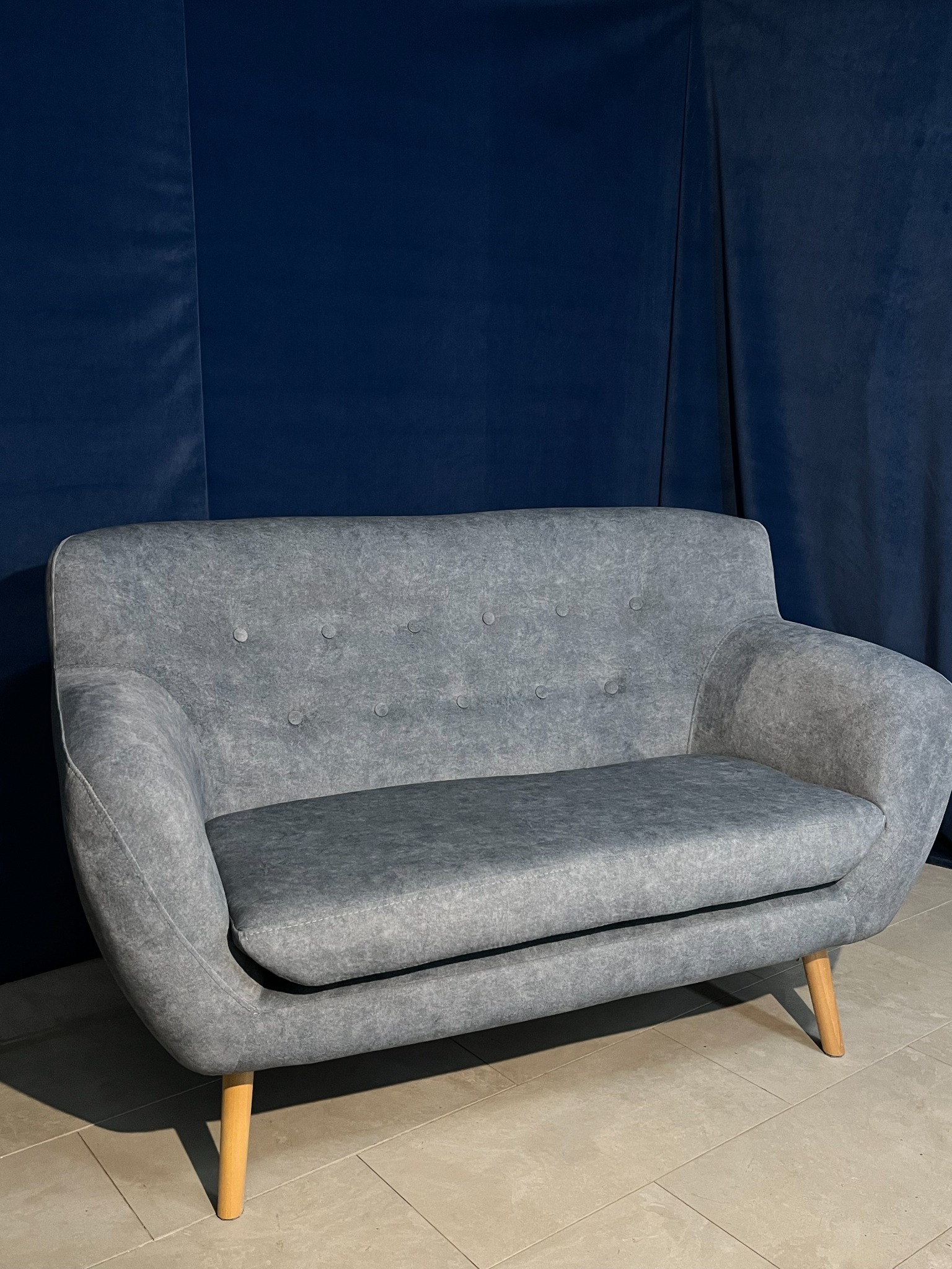 Szara sofa dwuosobowa w stylu retro na drewnianych nóżkach, z tapicerowanymi guzikami na oparciu, na tle ciemnoniebieskiej, aksamitnej zasłony.