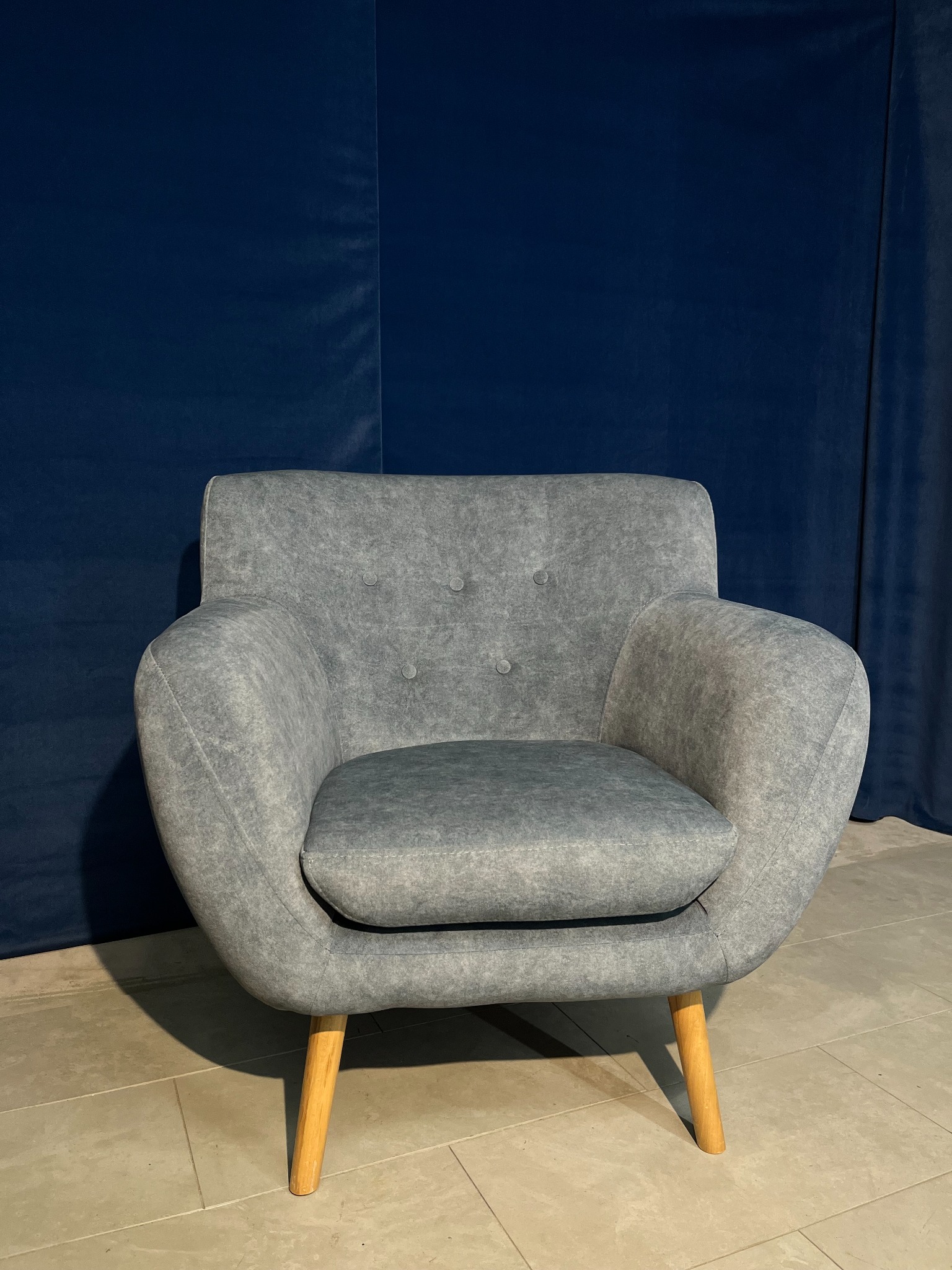 Stylowy, szary fotel tapicerowany z drewnianymi nóżkami na tle granatowej zasłony. Widoczne detale: przeszycia i guziki na oparciu. Minimalistyczny design.