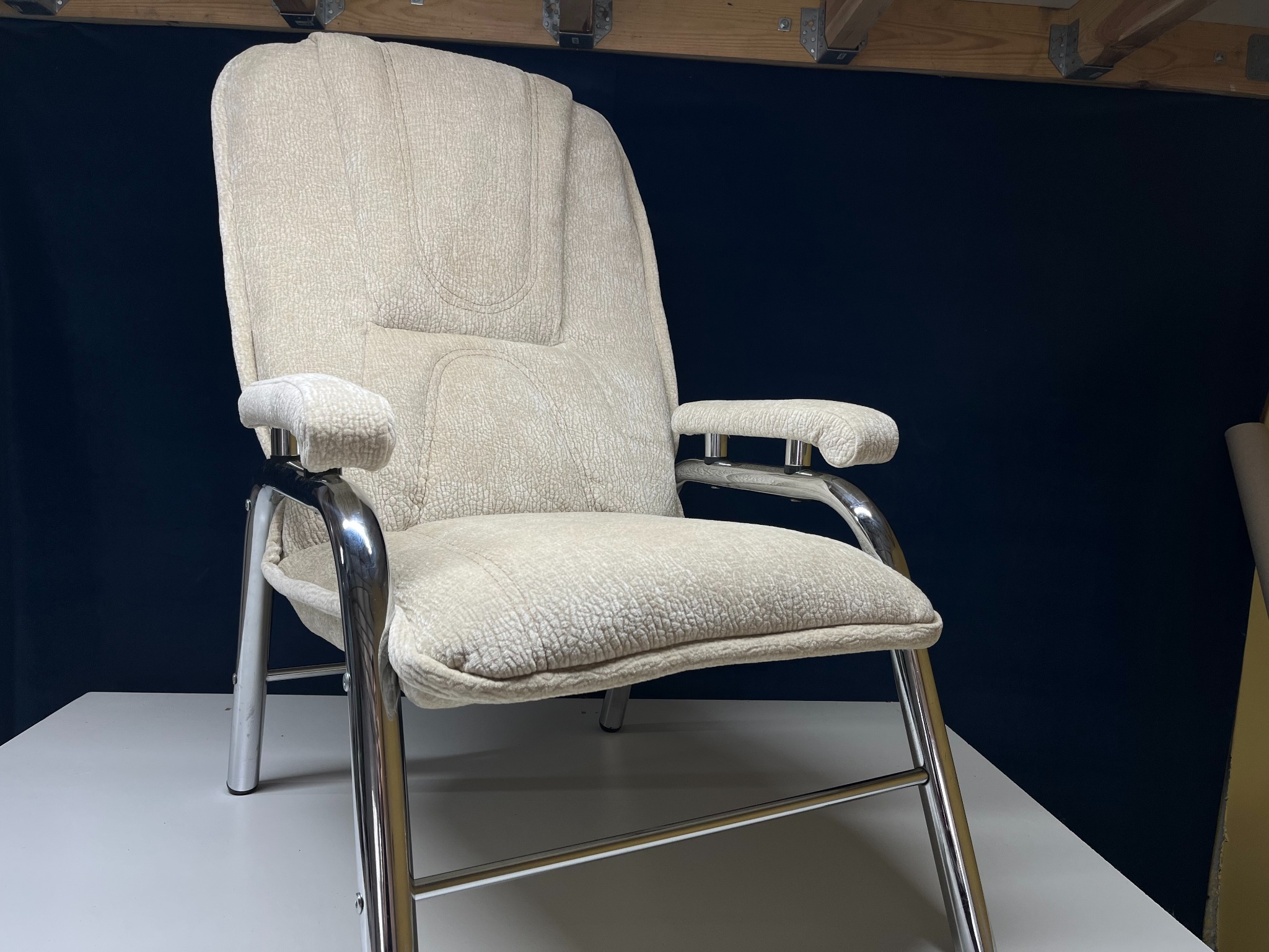 Beżowy fotel z tapicerką o wyraźnej fakturze i chromowanym stelażu, ustawiony na białym blacie na tle ciemnego tła. Widoczne elementy konstrukcji drewnianej.