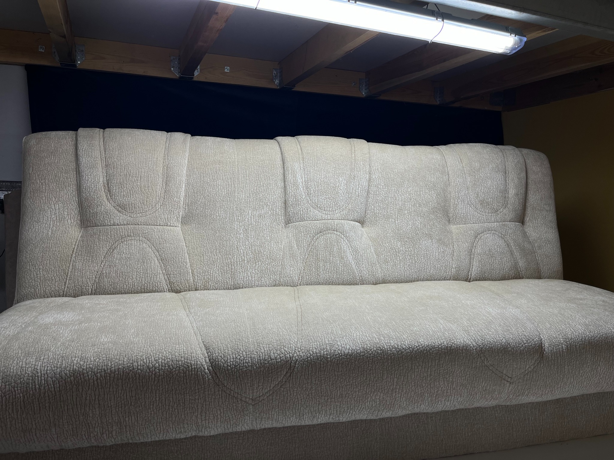 Jasna, tapicerowana sofa z wyraźną fakturą materiału, widoczna pod spodem konstrukcja sufitu i oświetlenie, ukazująca detale wykończenia.