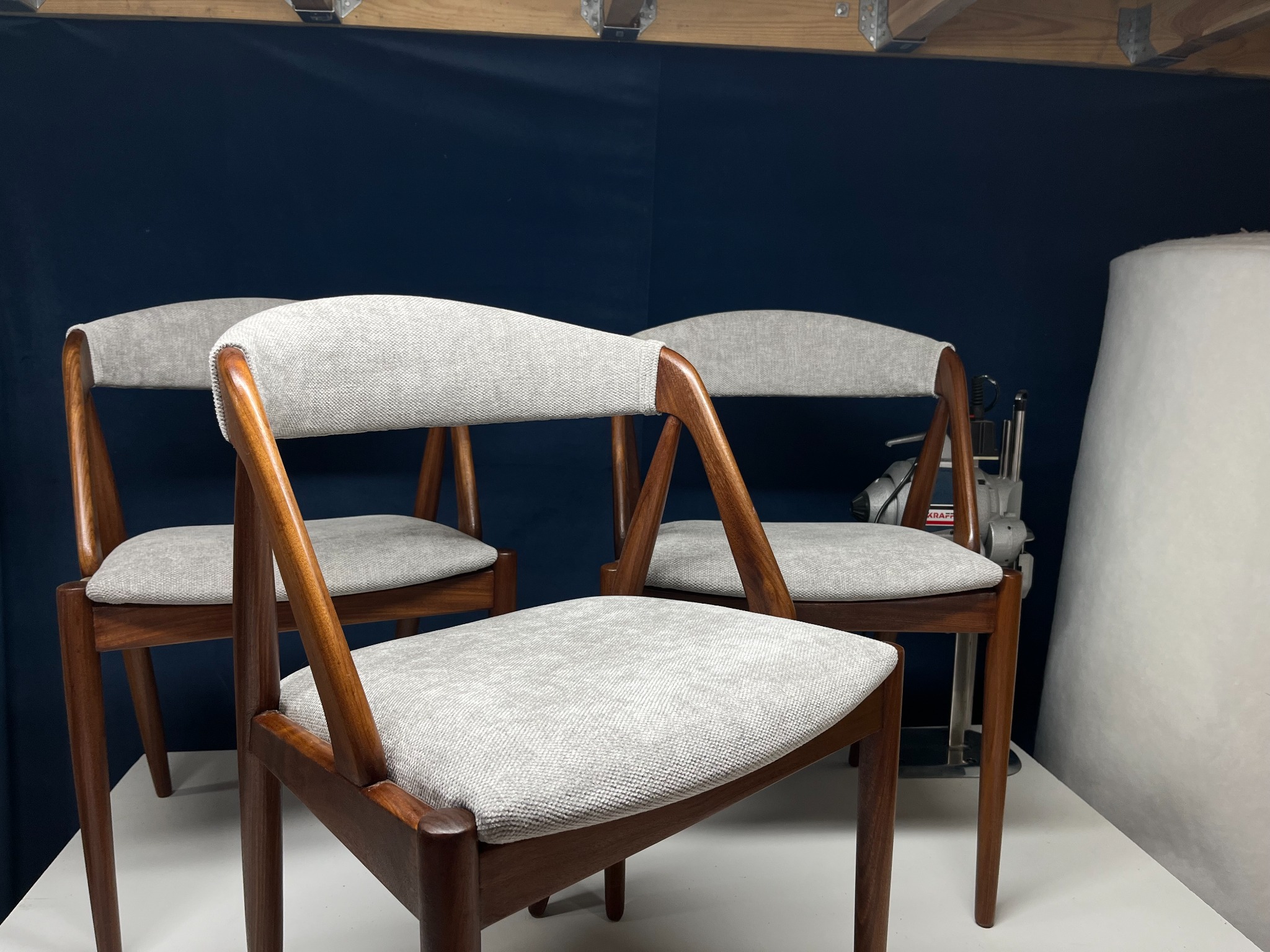 Trzy odnowione krzesła z drewnianą ramą i szarą tapicerką na siedziskach i oparciach, w tle narzędzie tapicerskie i rolka waty.